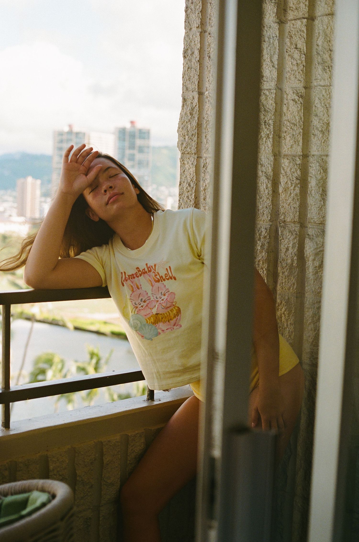 Woman in yellow graphic tee leaning against balcony doorway with city skyline view behind her.