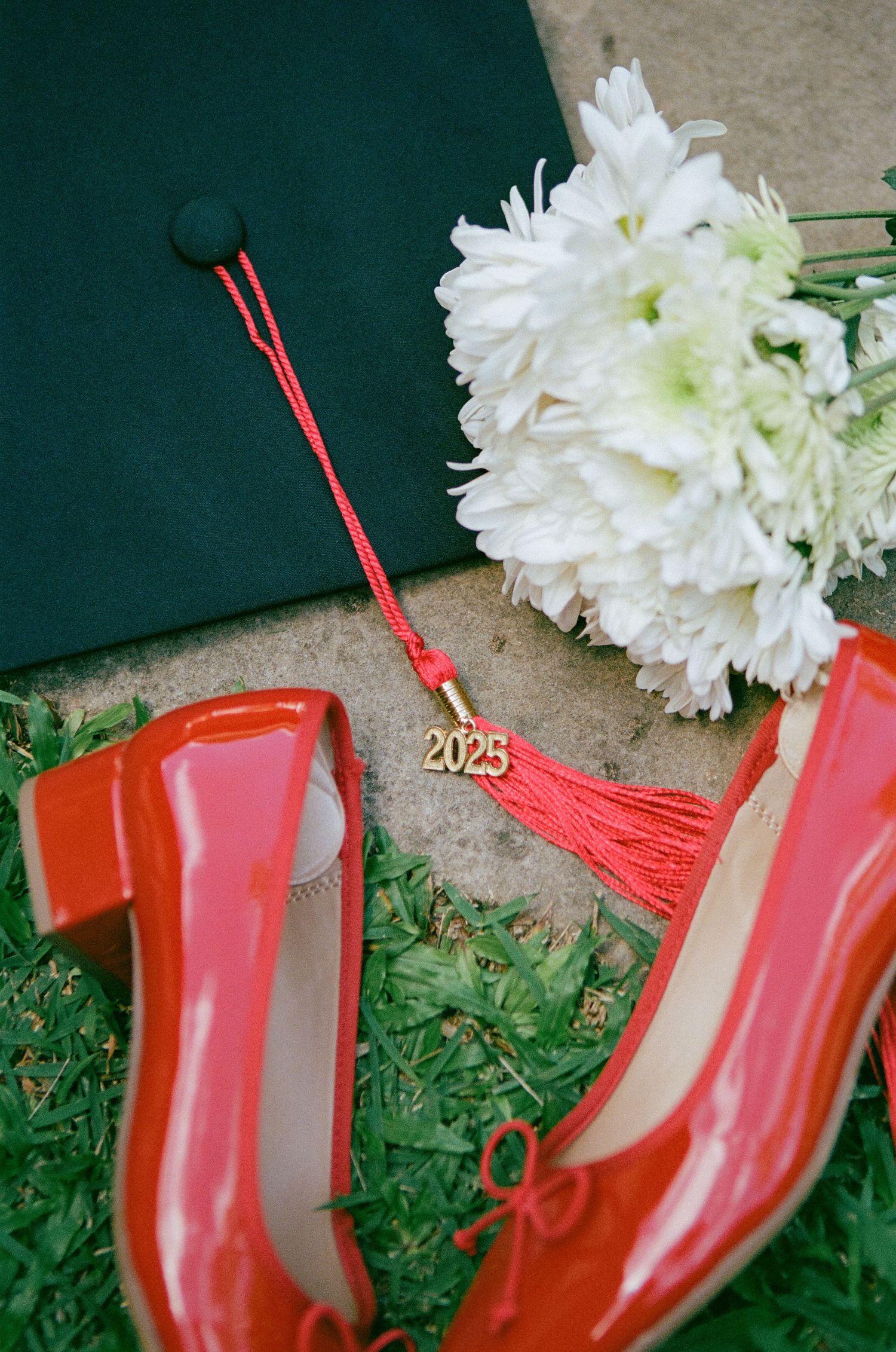 Class of 2025 graduation cap with red tassel beside red patent heels and white chrysanthemum bouquet.
