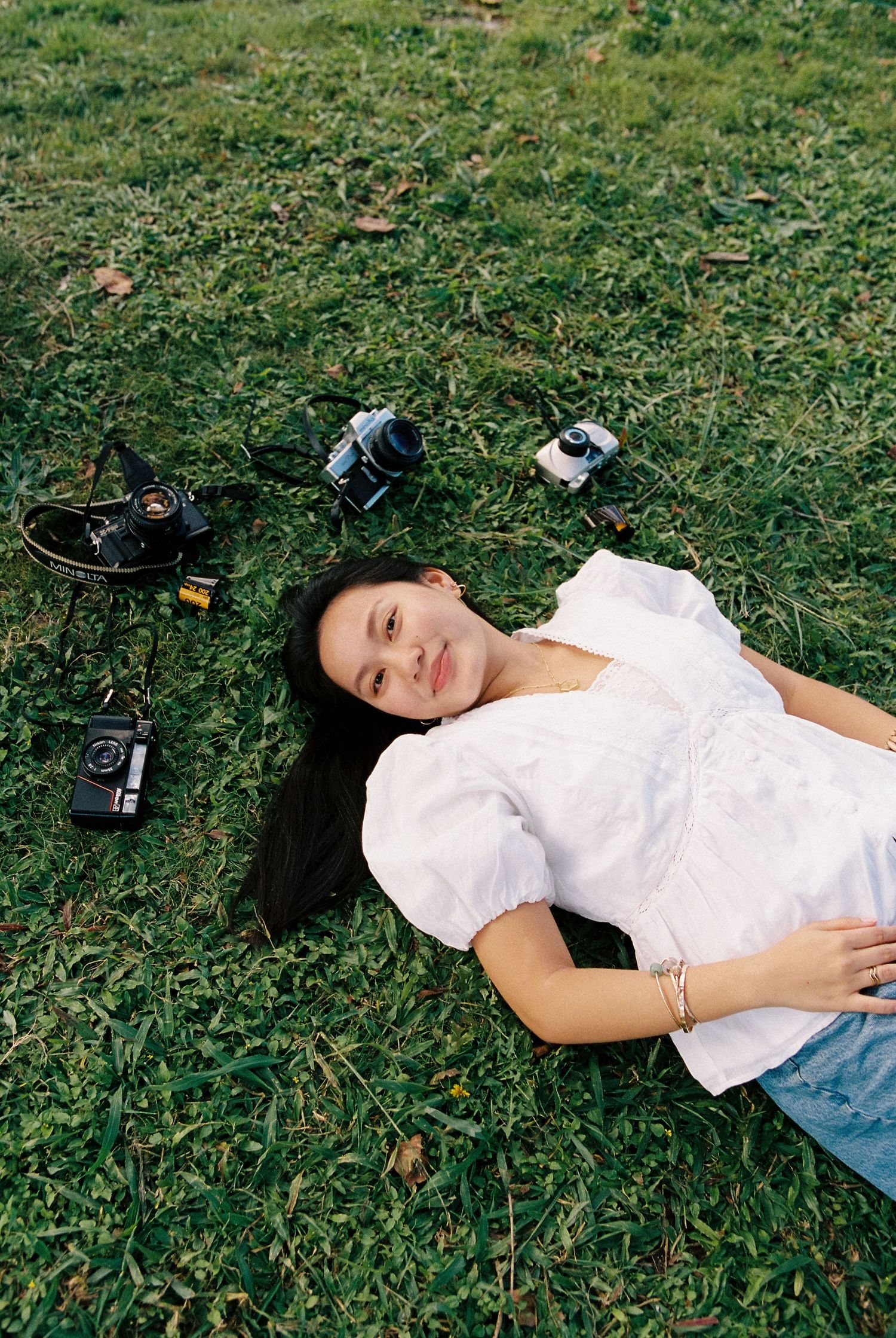 Young woman in white blouse lying on green grass surrounded by vintage film cameras, smiling up at camera.