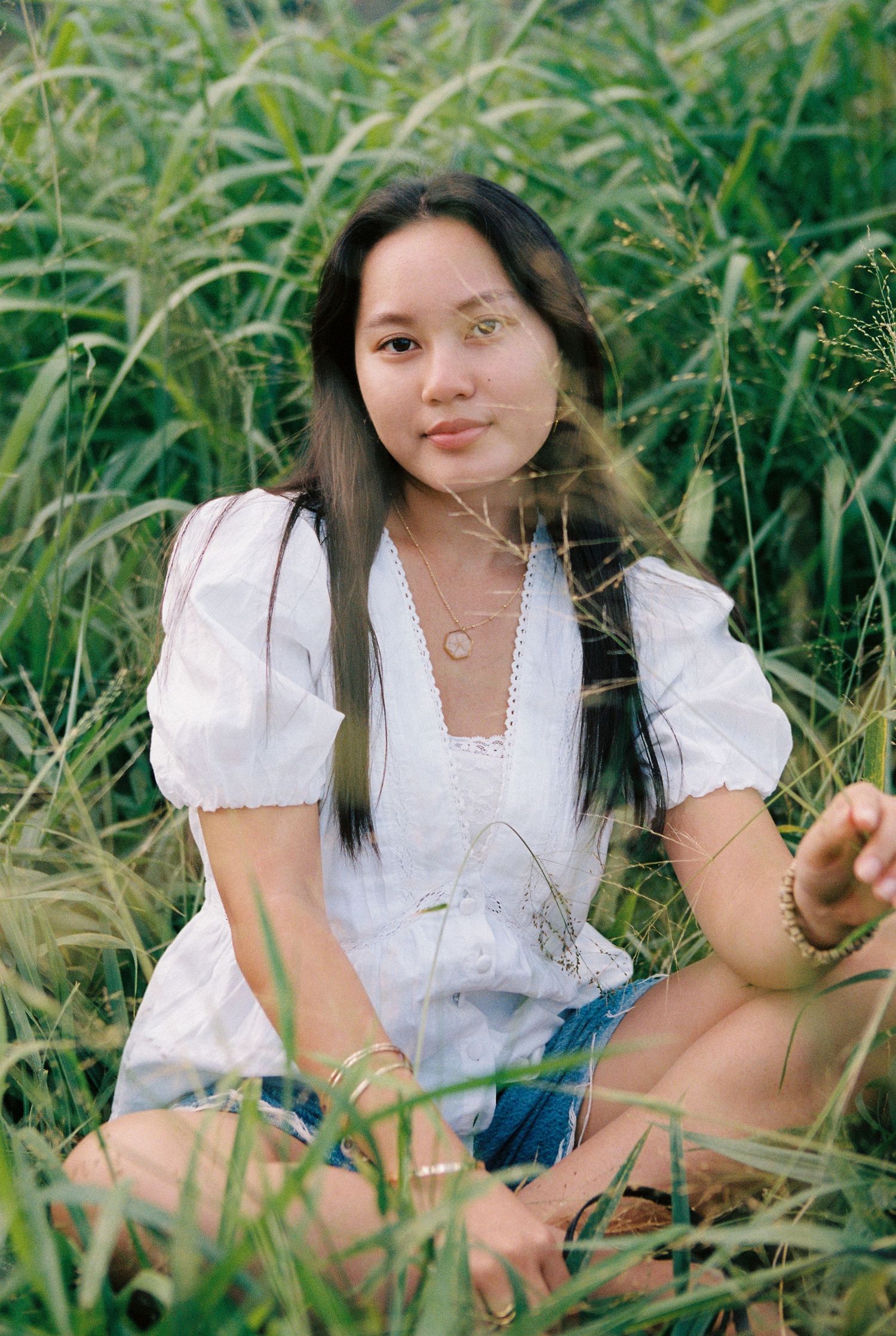 Young woman in white blouse sitting in tall green grass field, smiling at camera.