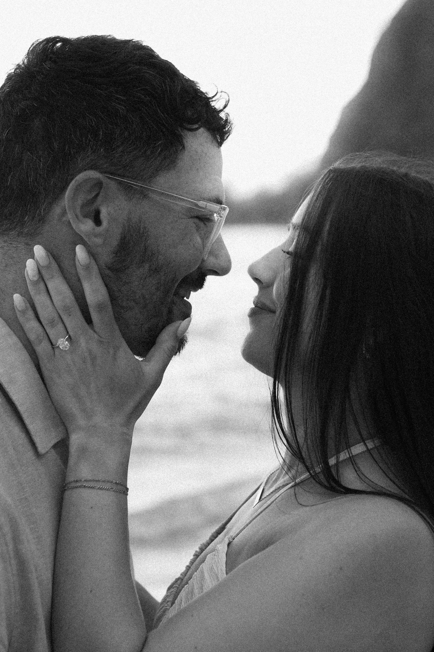 Black and white close-up of a laughing couple almost kissing on a beach, woman wearing engagement ring.