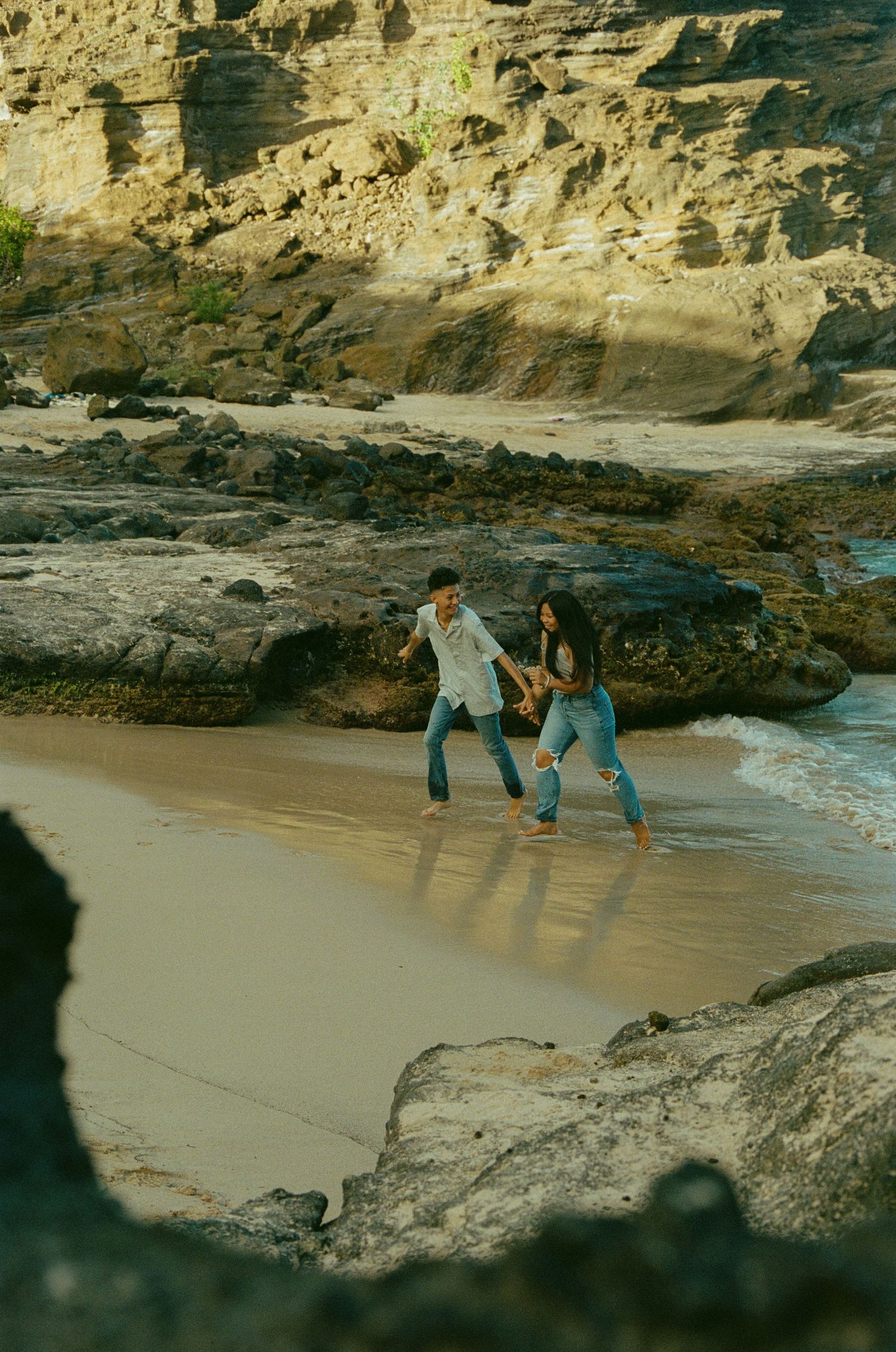 Couple laughs and runs together along a sandy beach framed by dramatic rocky cliffs.