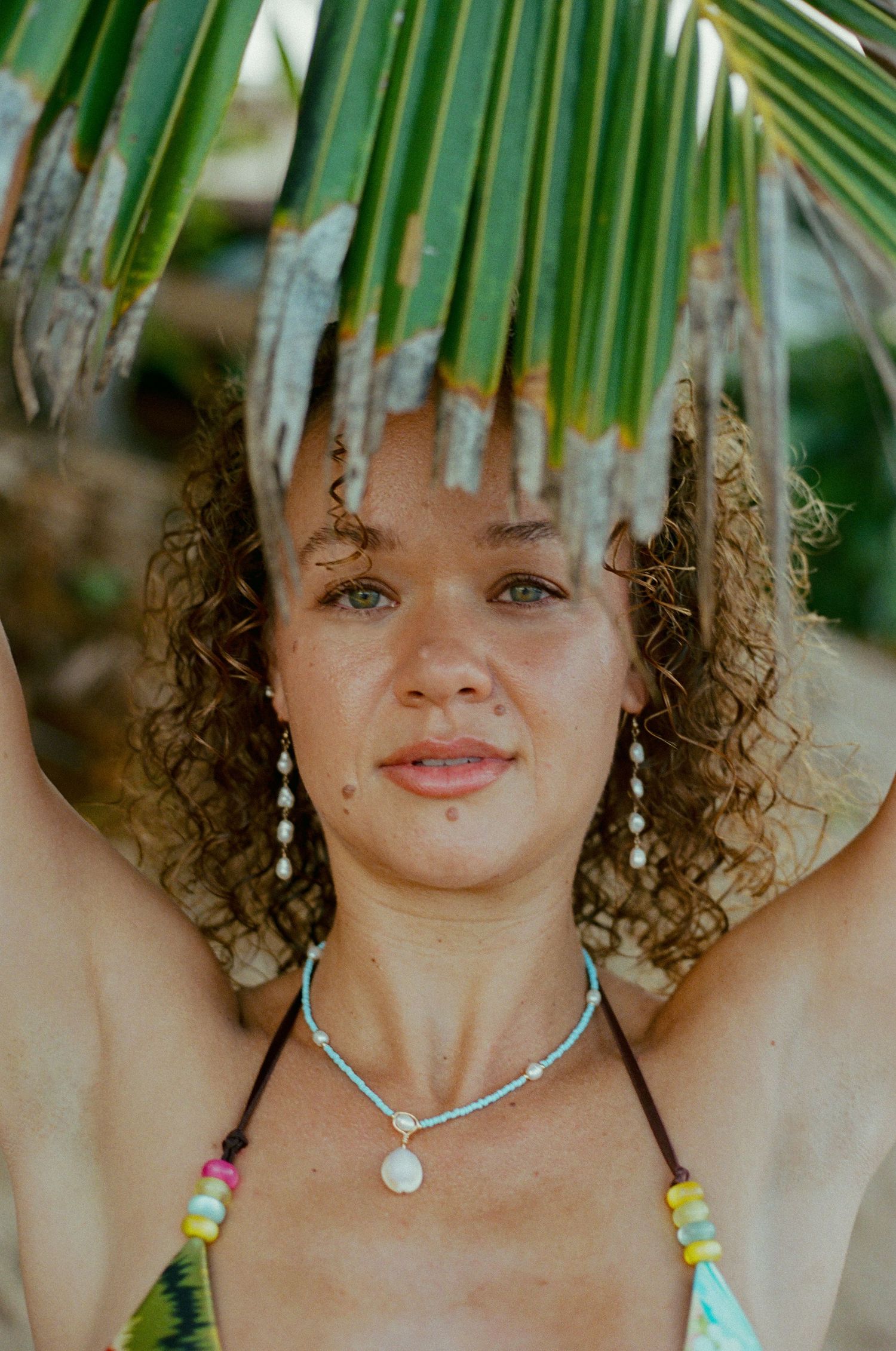 Close-up portrait of curly-haired woman holding palm leaf with colorful beaded necklace outdoors.