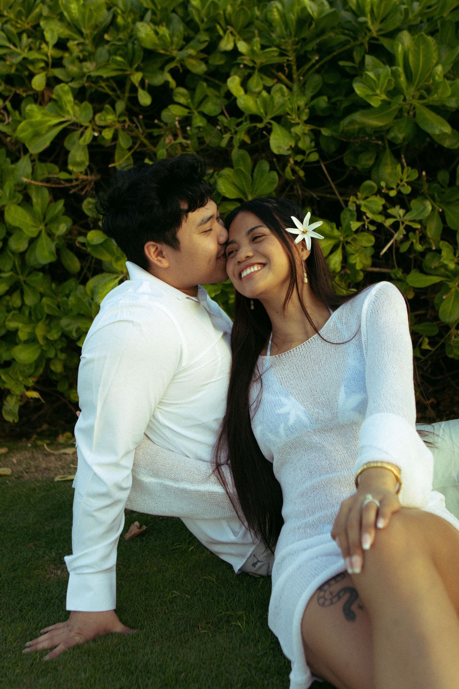 Couple in white outfits sharing a romantic kiss while sitting on grass with lush green foliage background.