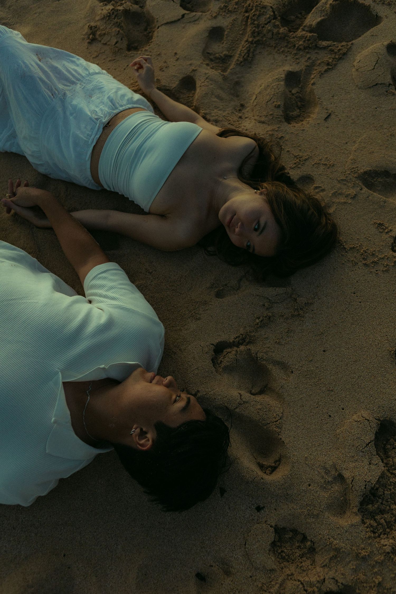 Couple lying head-to-head on sandy beach holding hands in a romantic overhead shot.