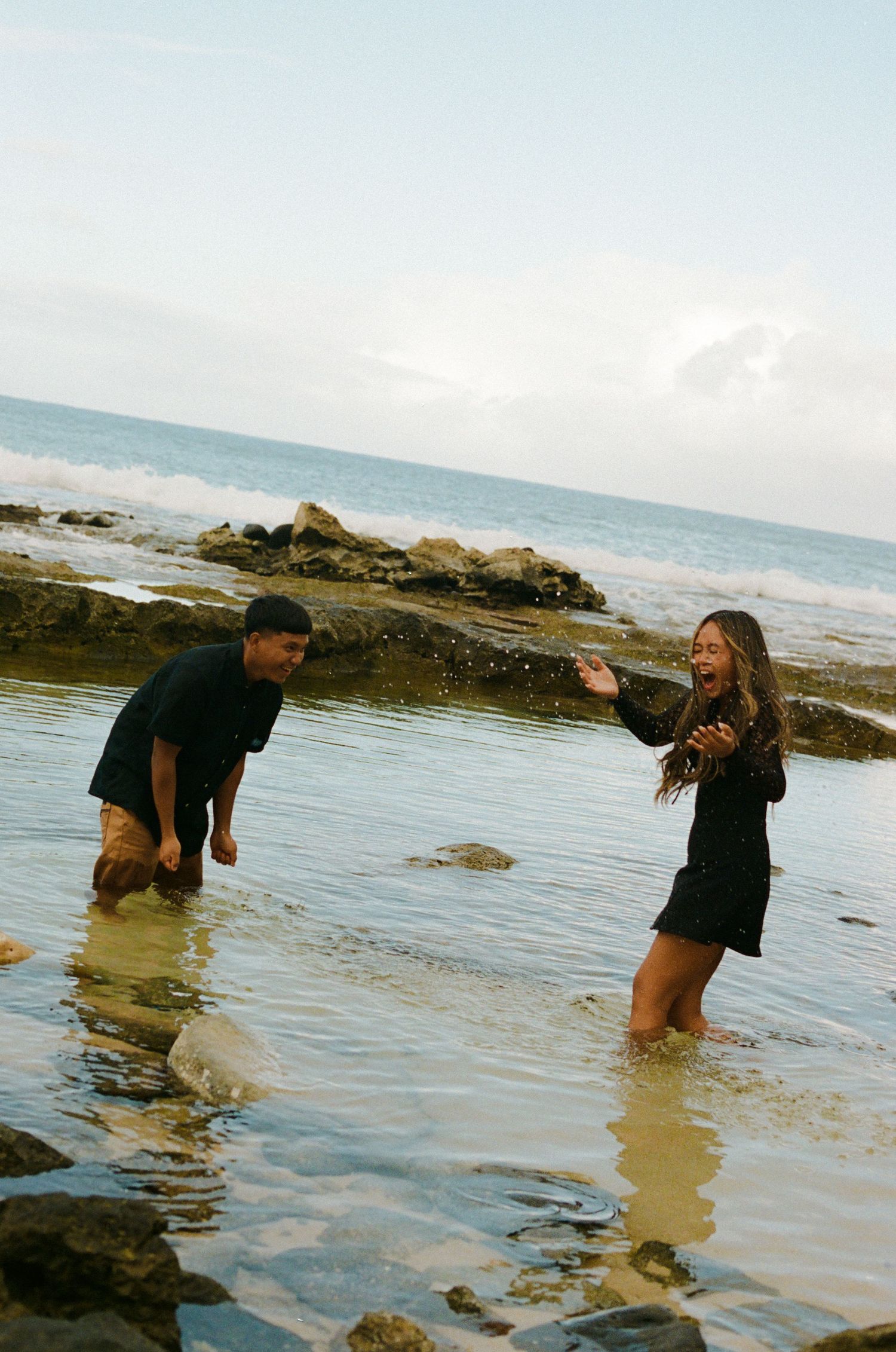 Couple laughing and splashing in a rocky ocean tide pool at the beach.