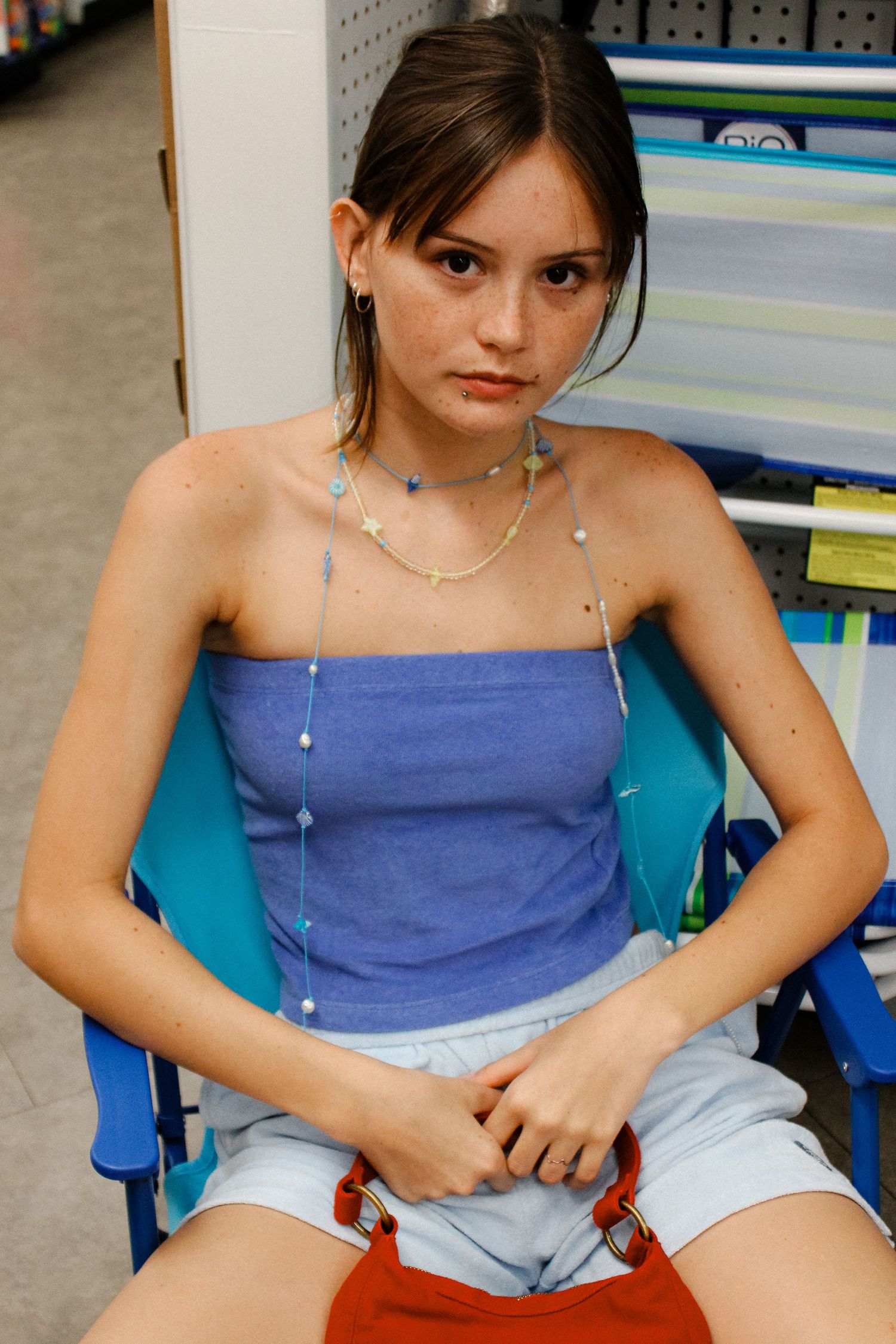 Young woman with freckles wearing blue tube top and layered necklaces sitting in a store.