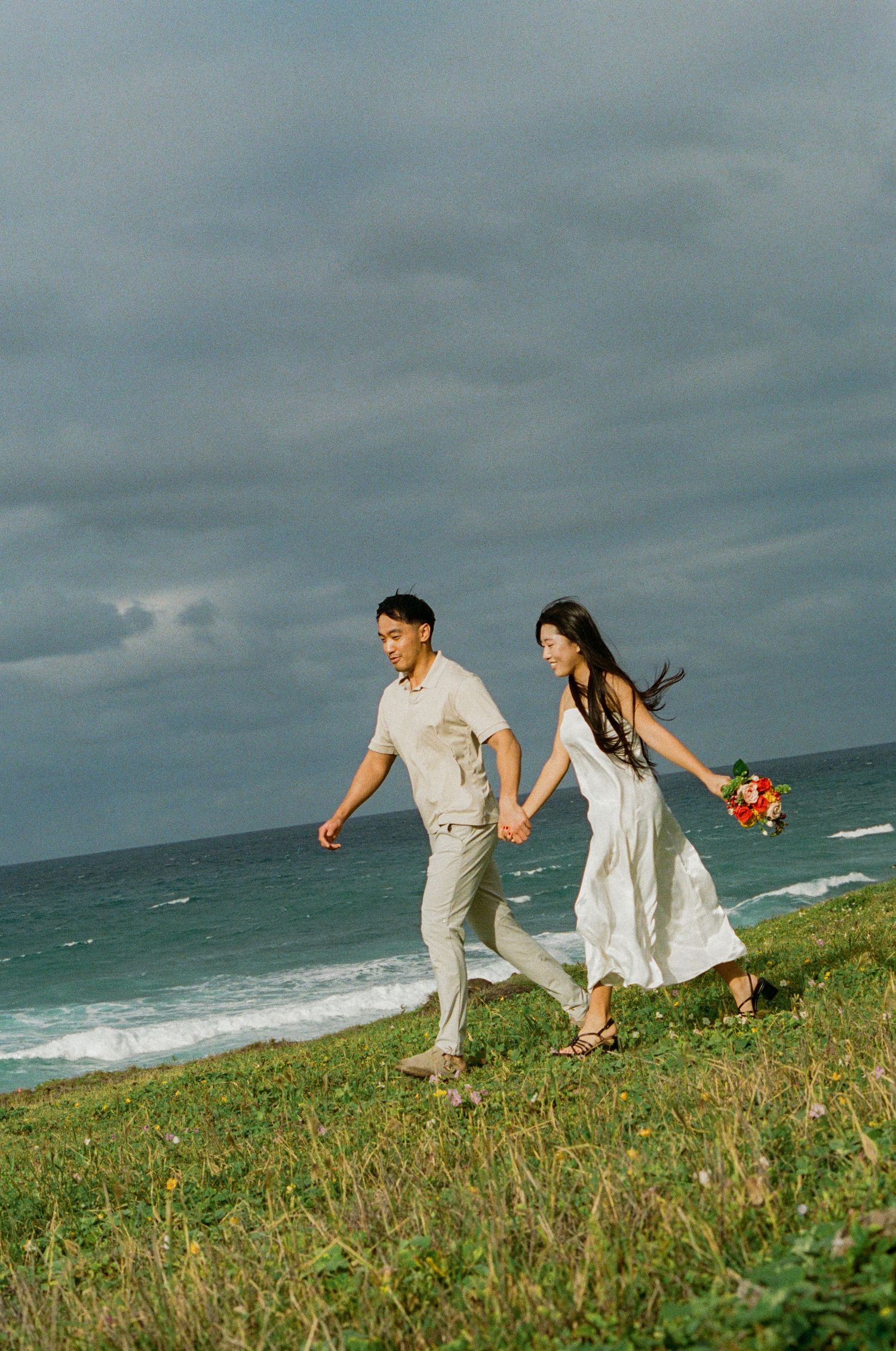 A couple walks hand-in-hand along a grassy coastal cliff with a bouquet and stormy ocean sky.