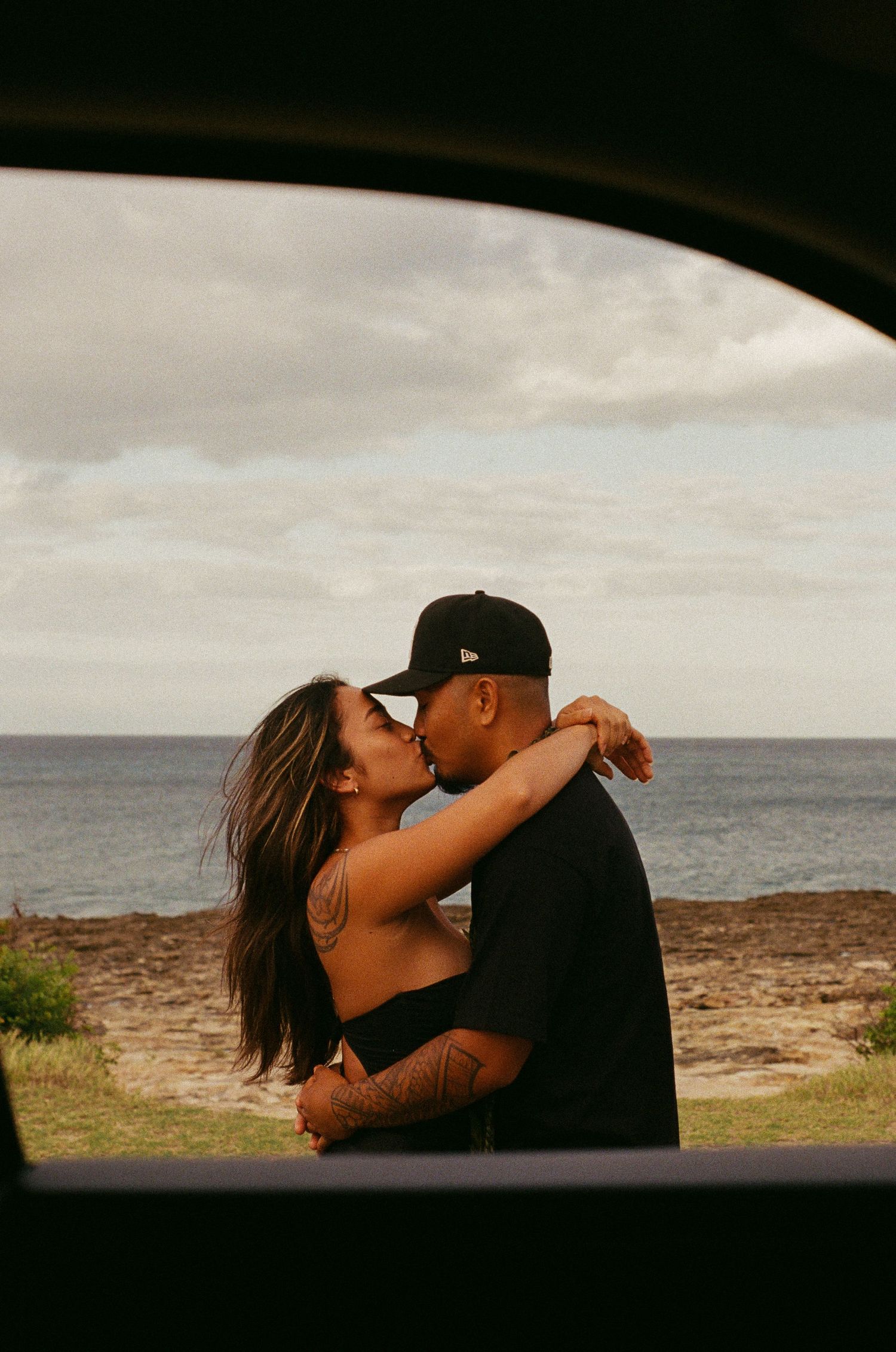 A couple shares a passionate kiss by the ocean, framed through a car window opening.