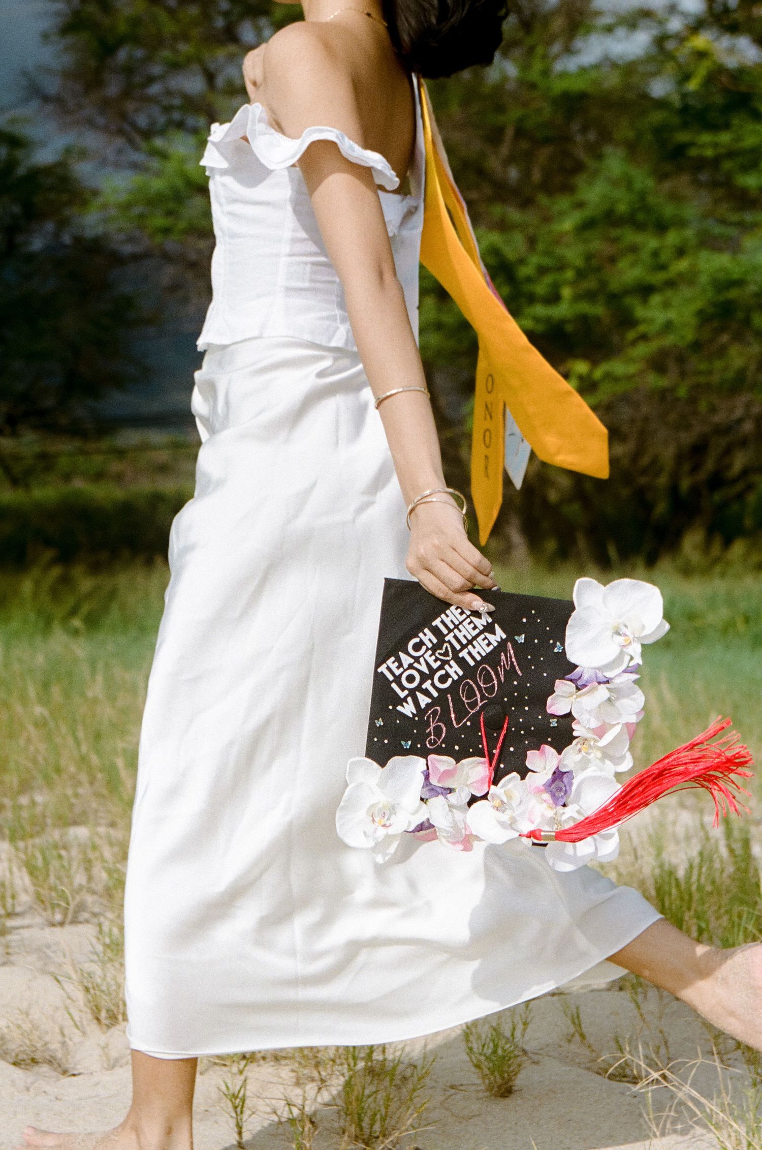 Graduate in white ruffled dress carrying floral-decorated mortarboard reading Teach them to bloom.