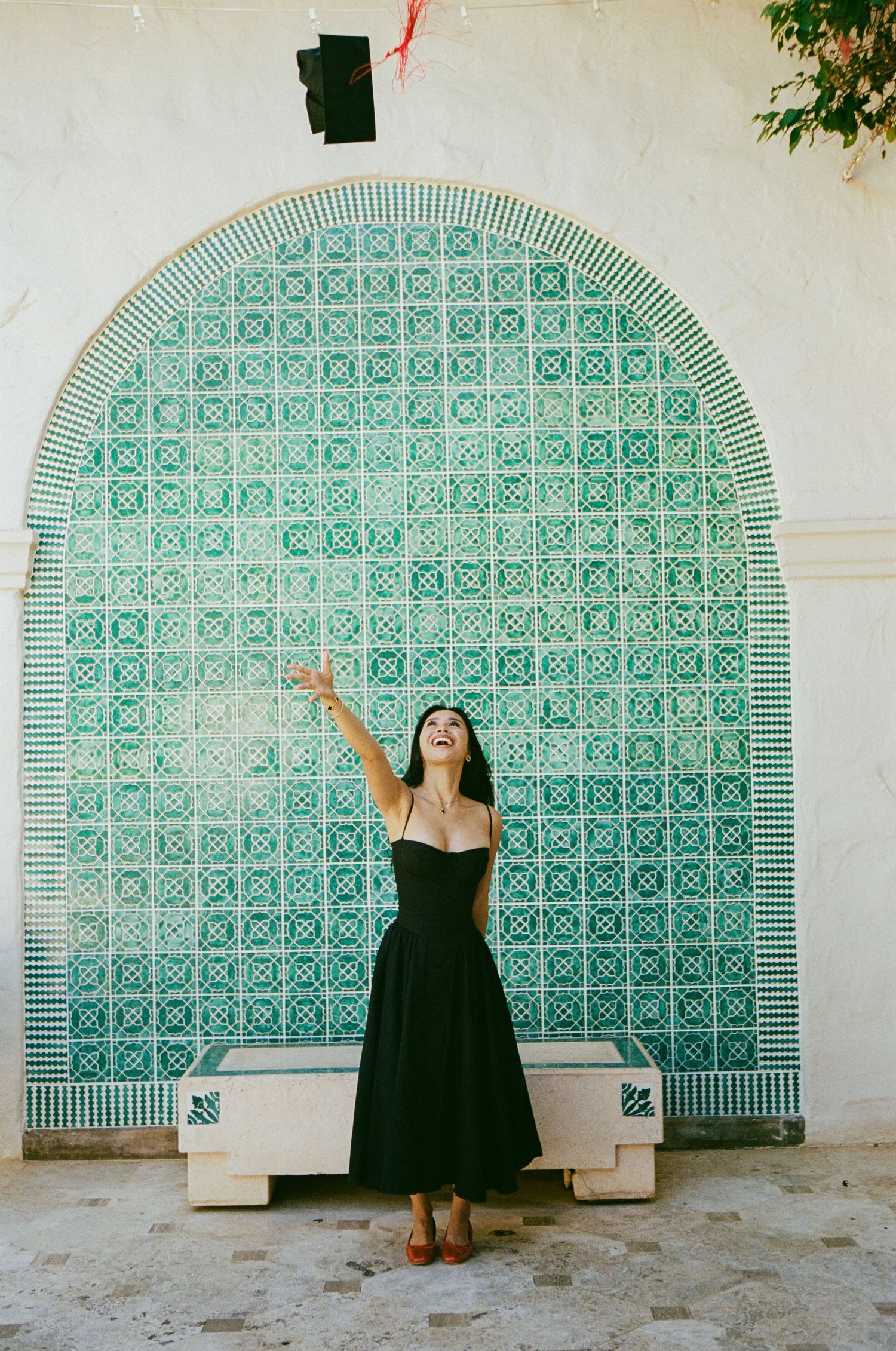 Laughing woman in black dress tossing graduation cap against decorative green tiled arch wall.