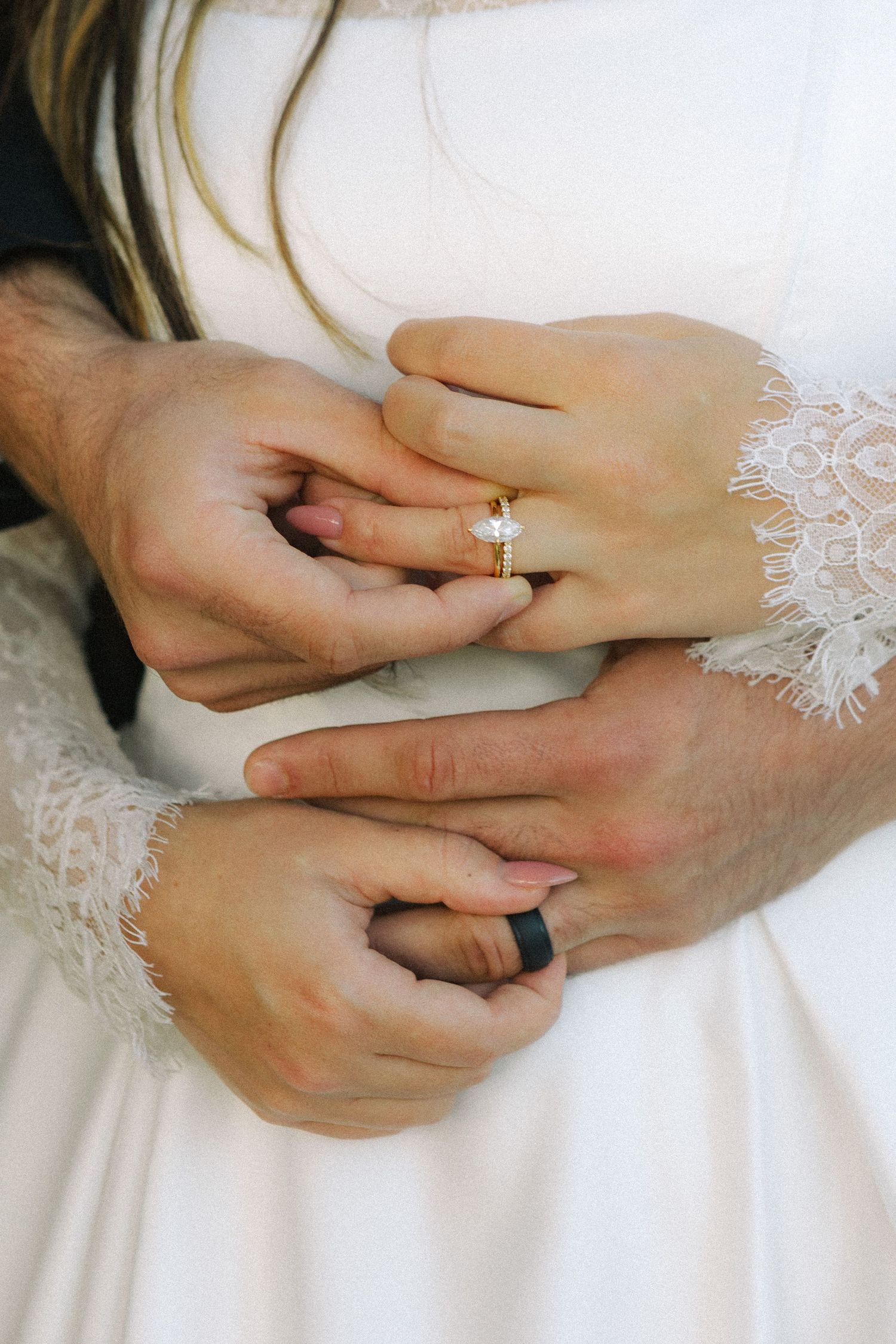 Close-up of newlyweds hands, bride wearing gold diamond ring with lace sleeve, groom with black wedding band.