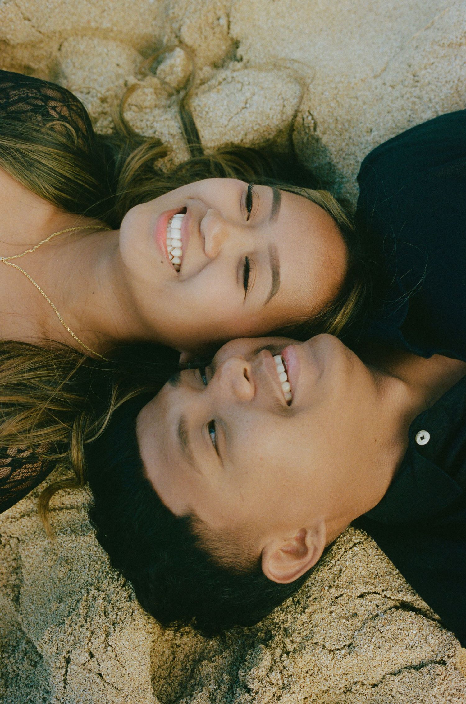 A happy couple lies head-to-head on sandy ground, smiling and laughing together closely.