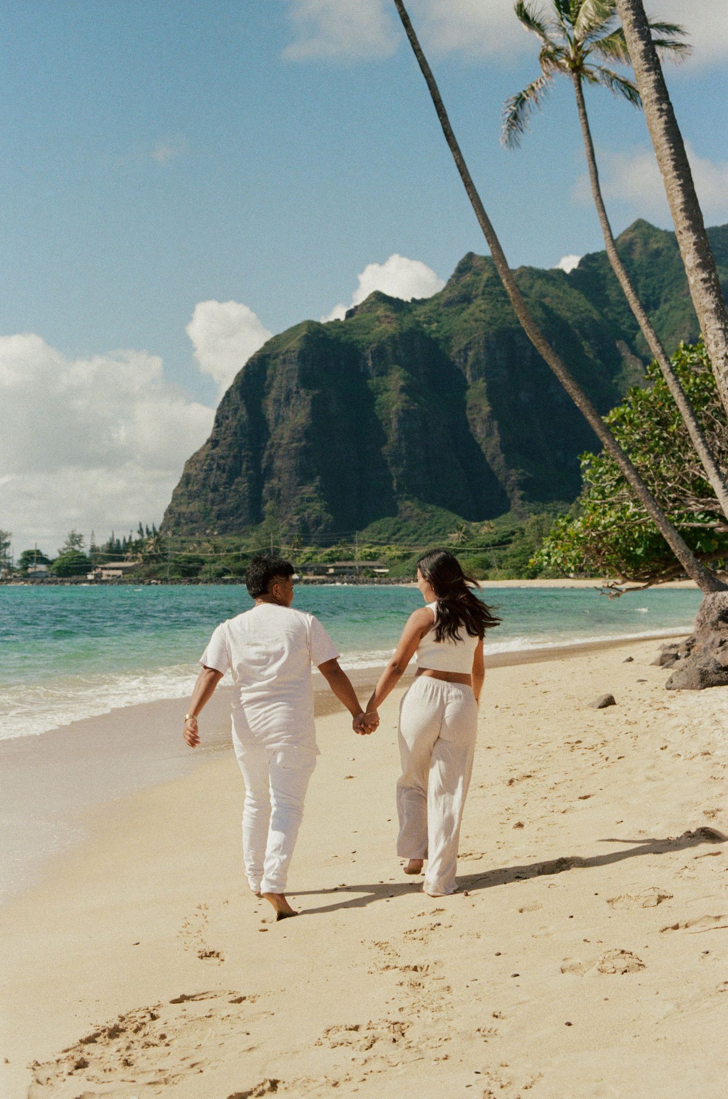 A couple in matching white outfits walks hand-in-hand along a tropical beach with palm trees.