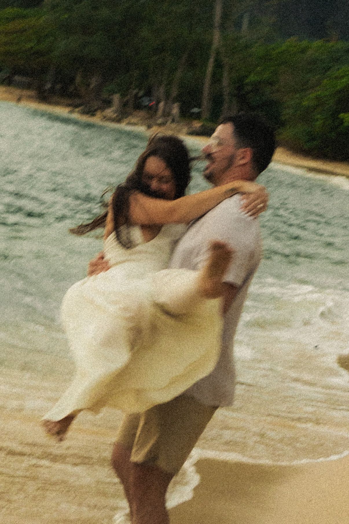 Couple embracing and spinning on tropical beach at dusk in white and gray outfits.