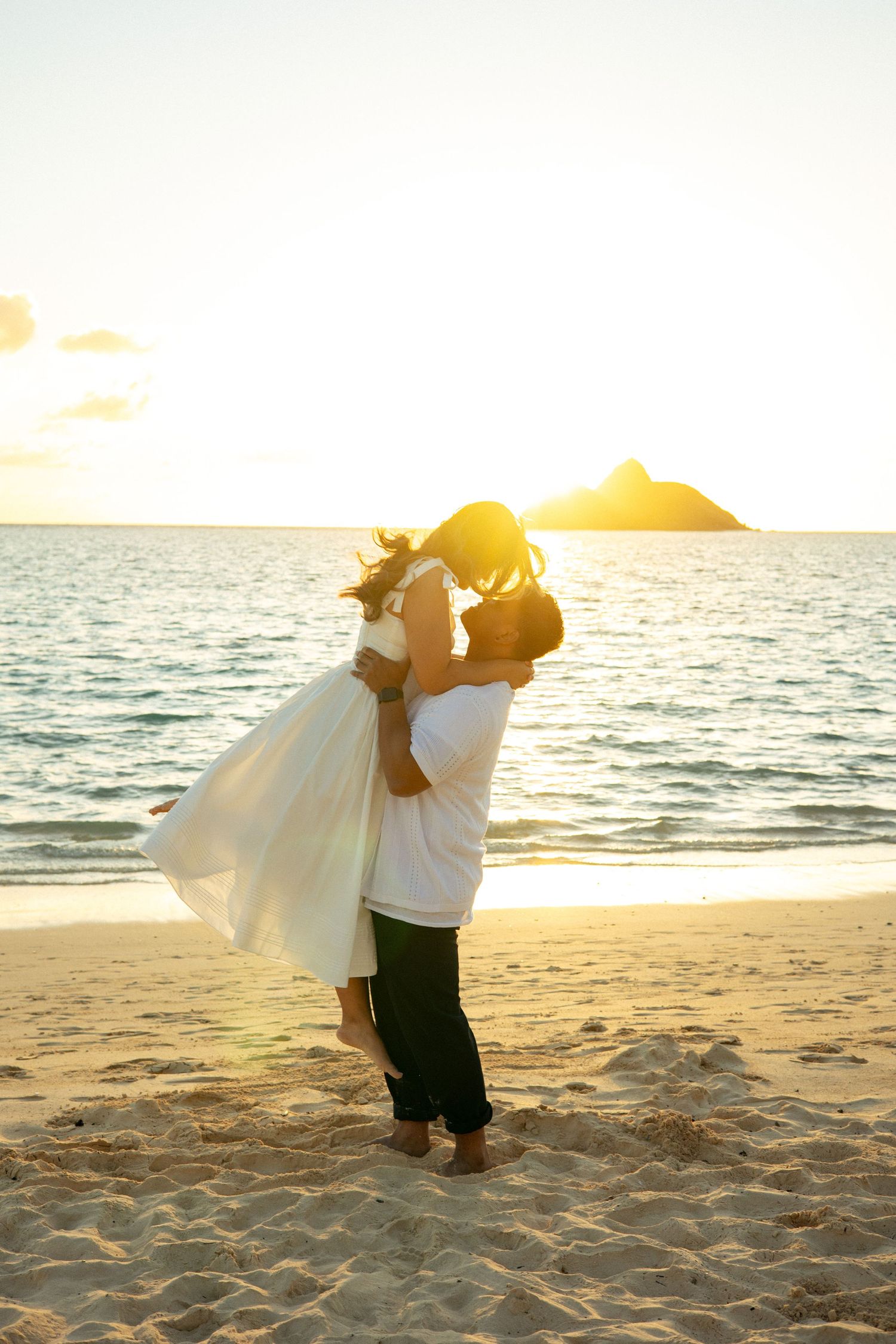 Man lifting woman in white dress during romantic beach sunset with island silhouettes in background.