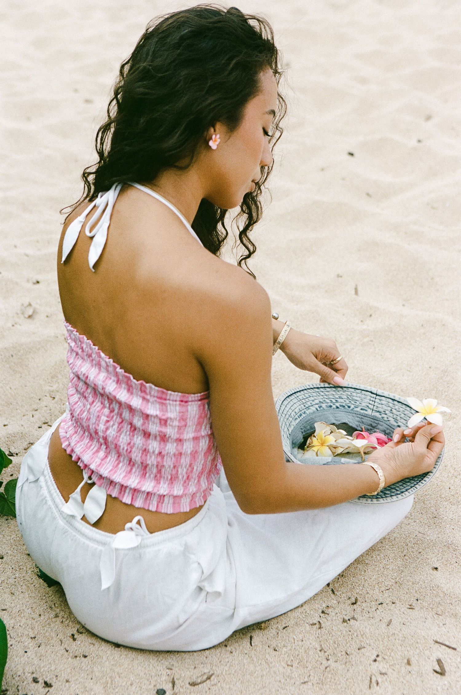 Woman in pink smocked crop top and white pants sitting on sandy beach holding a hat with flowers.