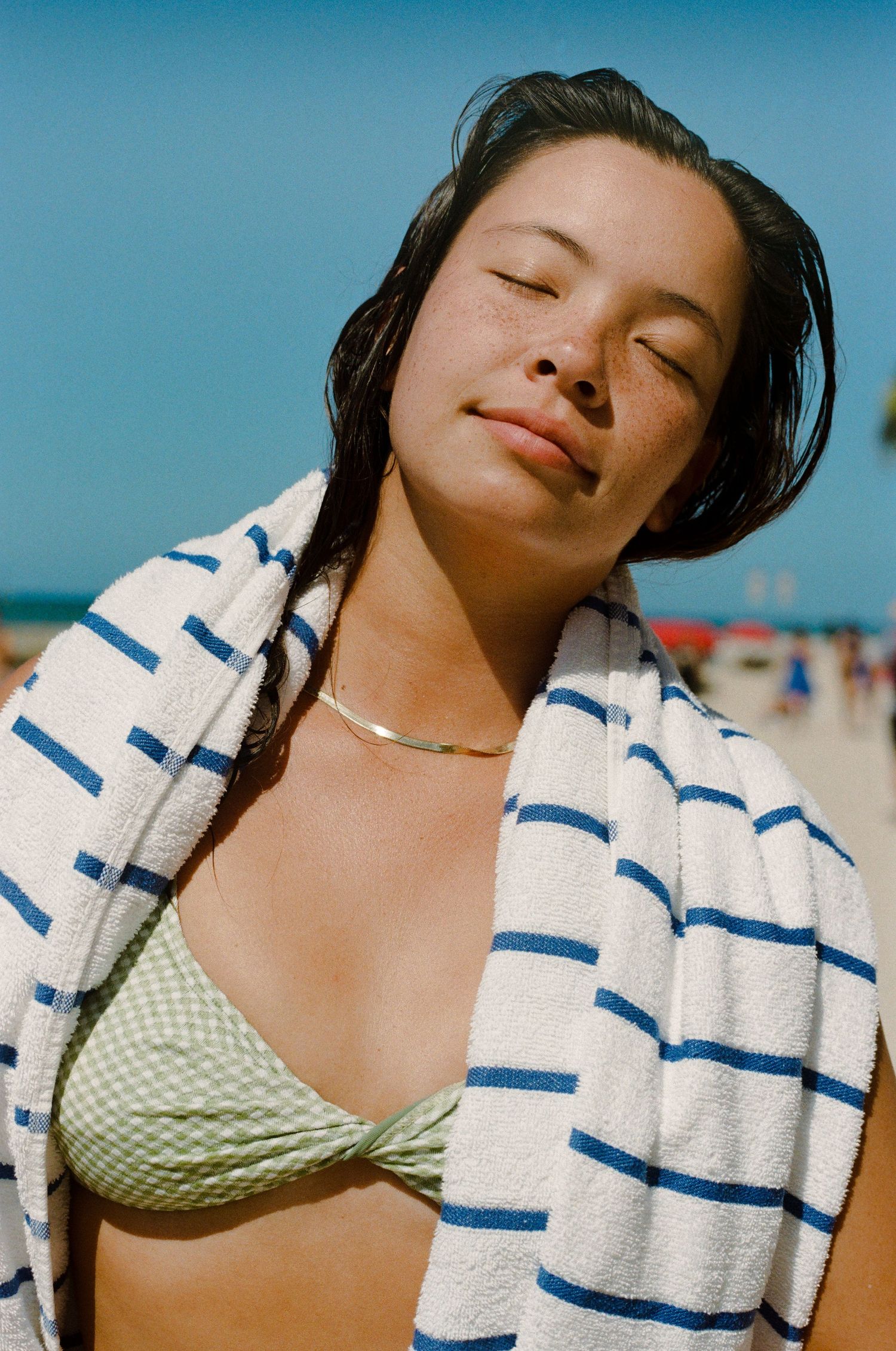 Woman wearing striped towel and green bikini top with gold necklace relaxing on sunny beach.