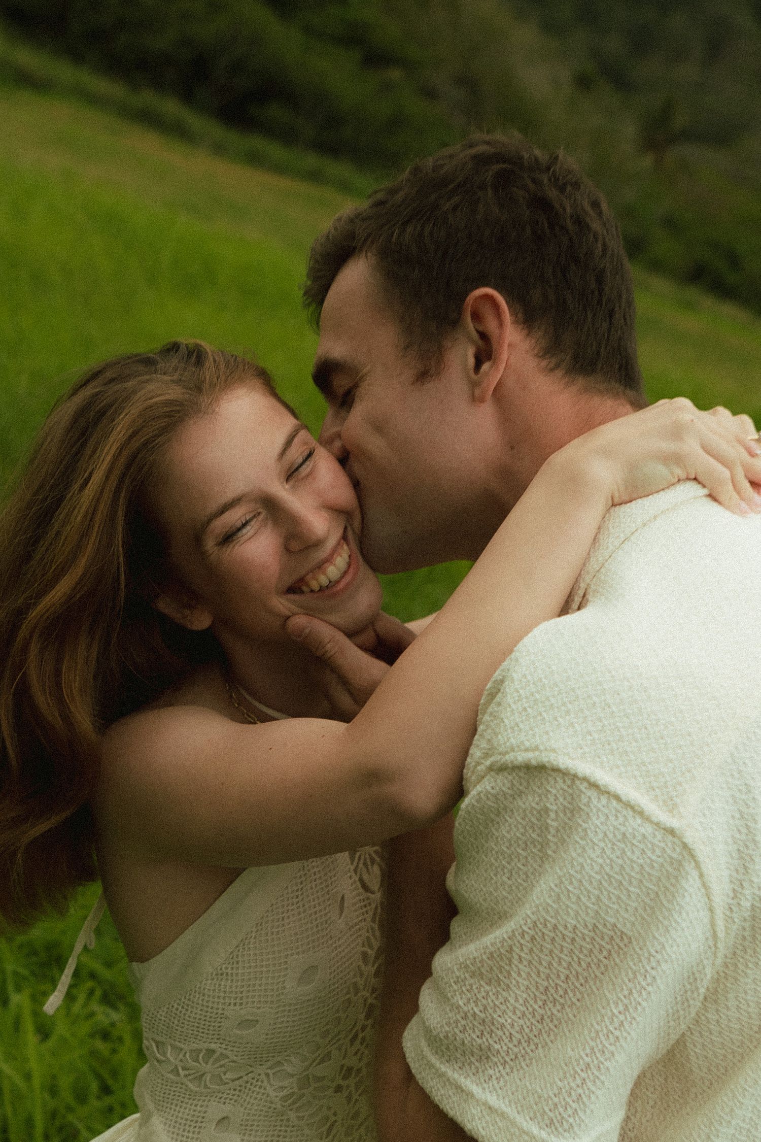 Man kissing smiling woman on cheek in lush green Hawaiian valley, both wearing white.
