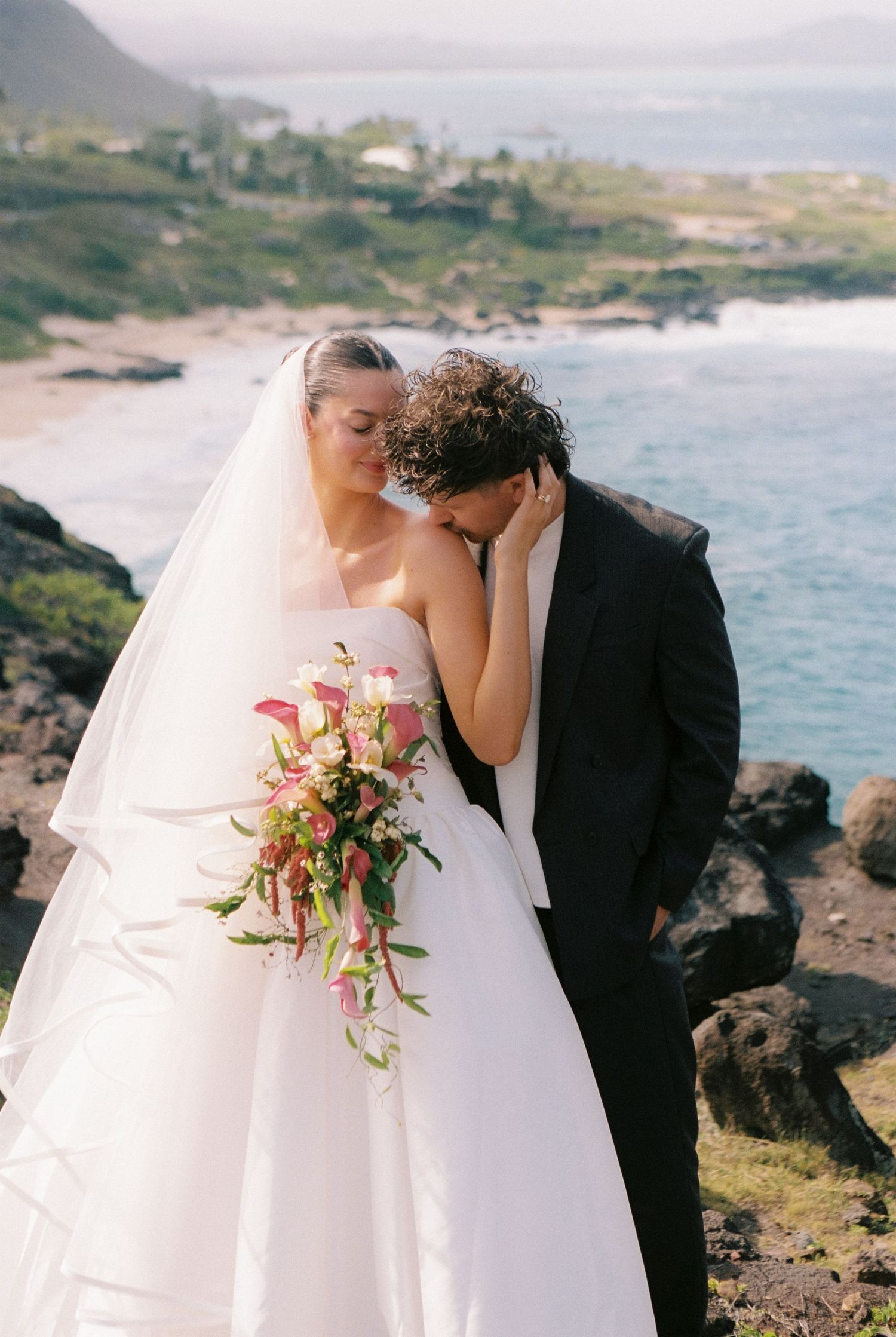 Bride and groom share romantic moment on rocky Hawaiian coastline, bride holding pink tropical bouquet in white gown.