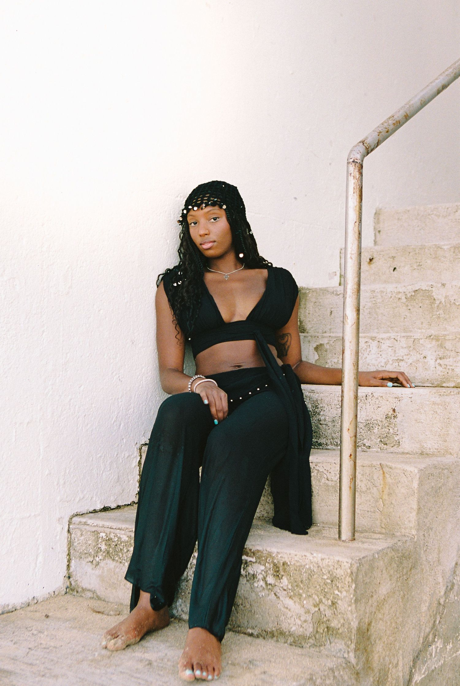 Young Black woman in black crop top and wide-leg pants sitting on stone steps barefoot.