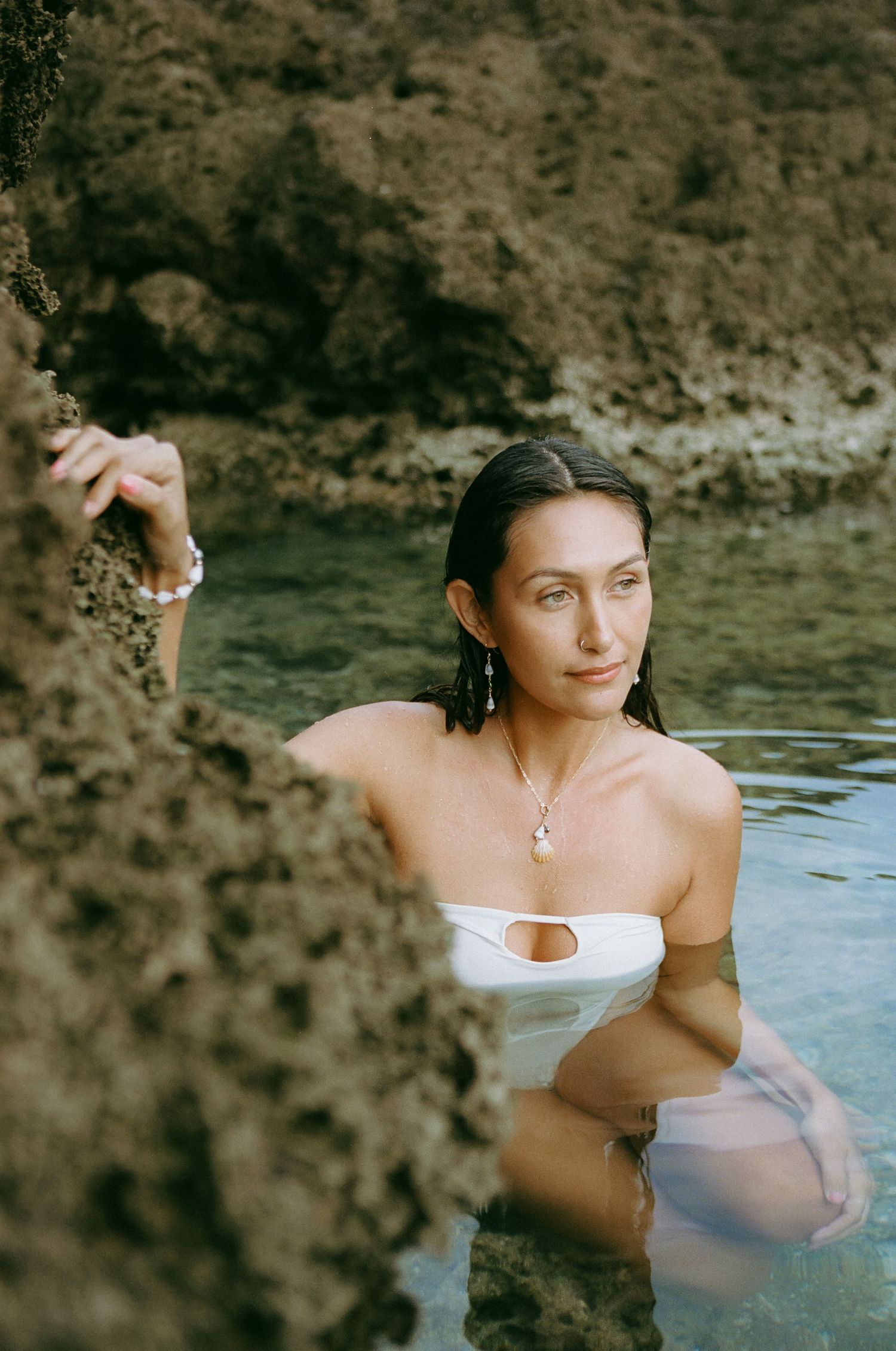Woman in white bandeau swimsuit wearing pearl jewelry standing in natural ocean rock pool.