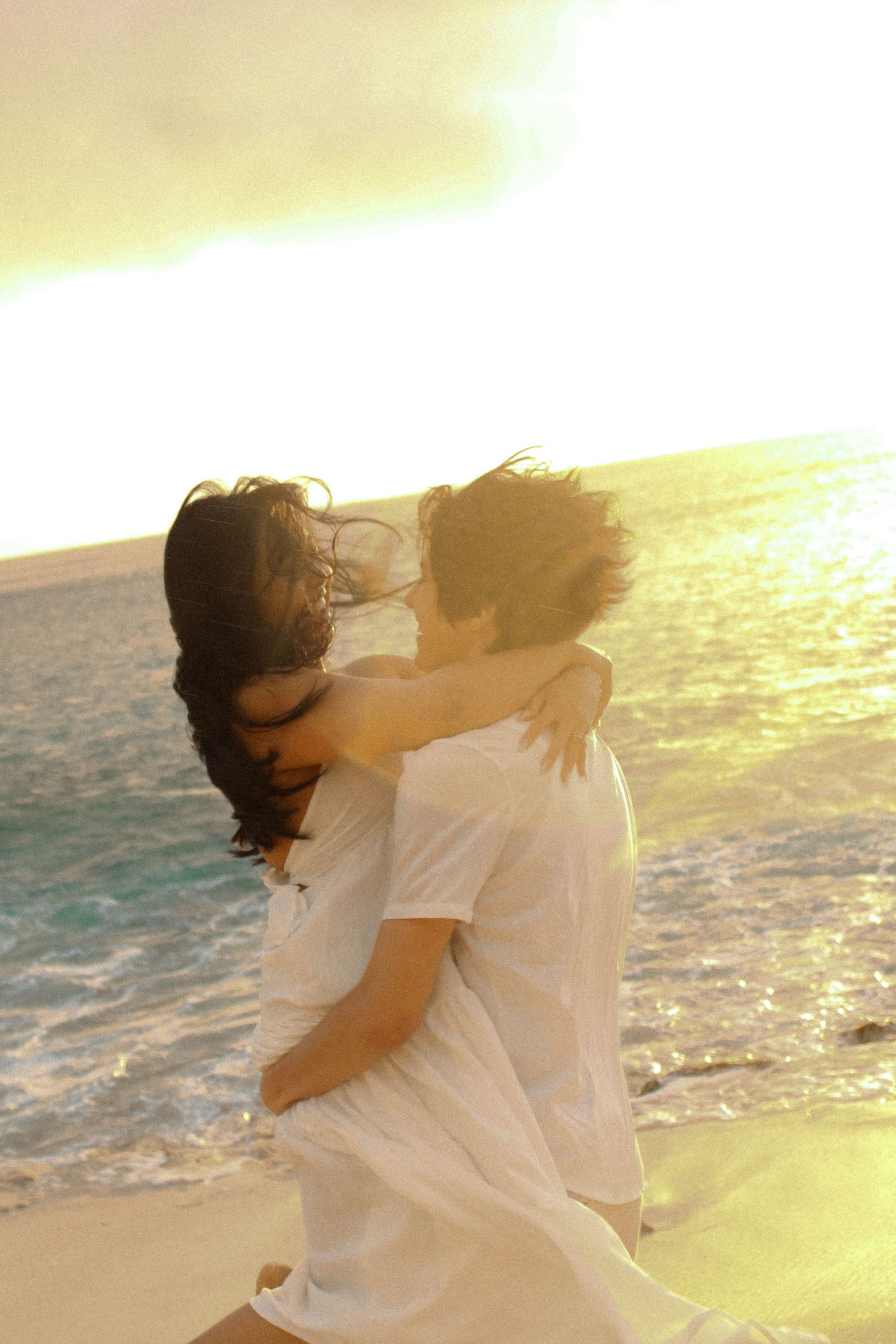 Romantic couple embracing on a bright sunlit beach with ocean waves and golden light behind them.