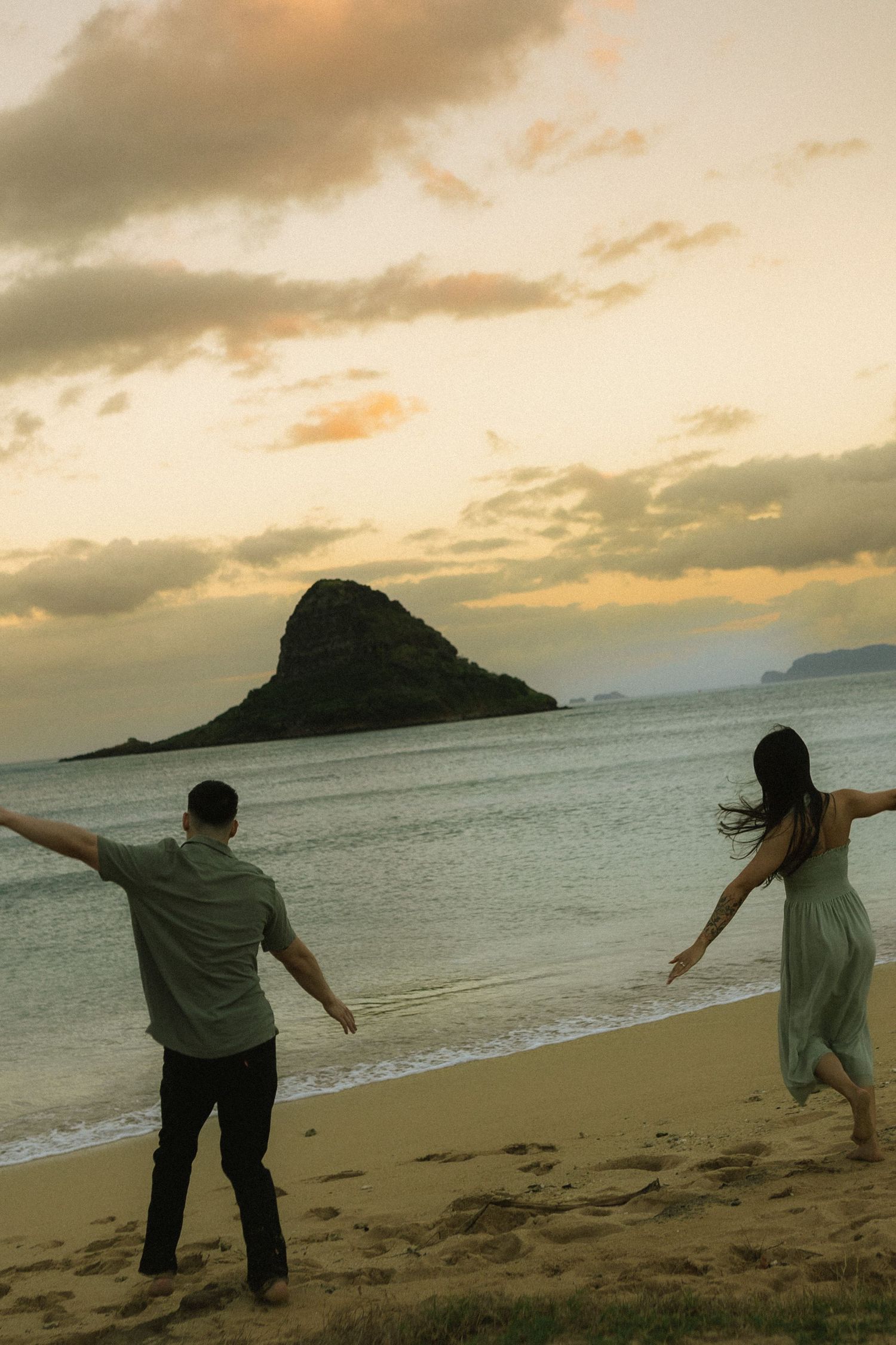 Couple running playfully toward the ocean at sunset with a dramatic island silhouette.