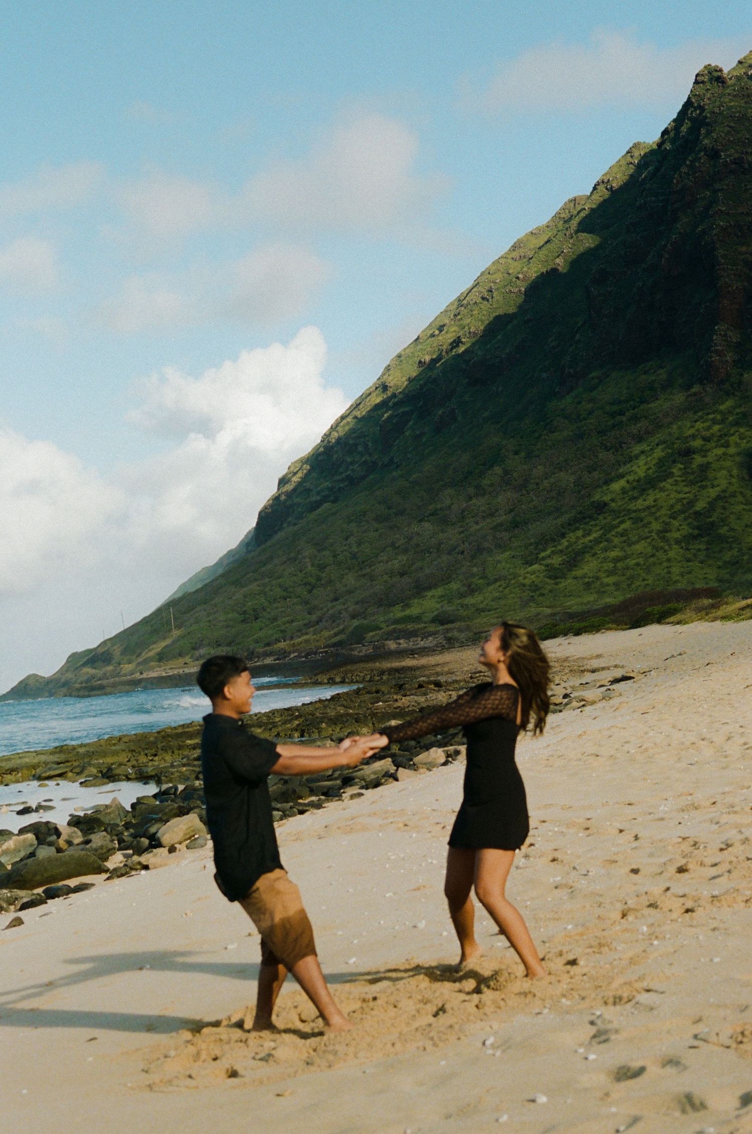 Couple holding hands and dancing playfully on a scenic Hawaiian beach with green mountains.