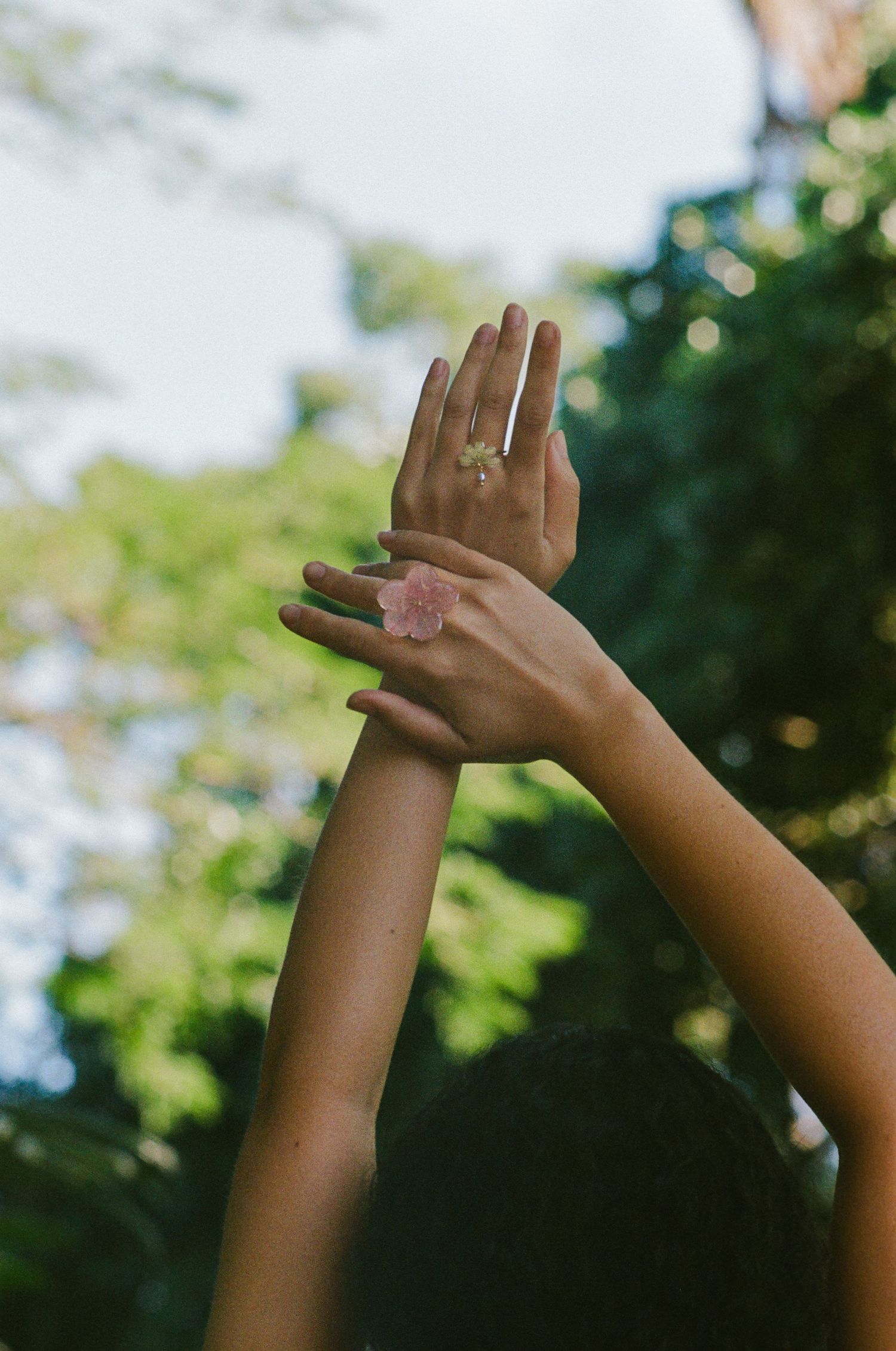 Hands raised toward sky wearing a pink flower ring with green trees in background.