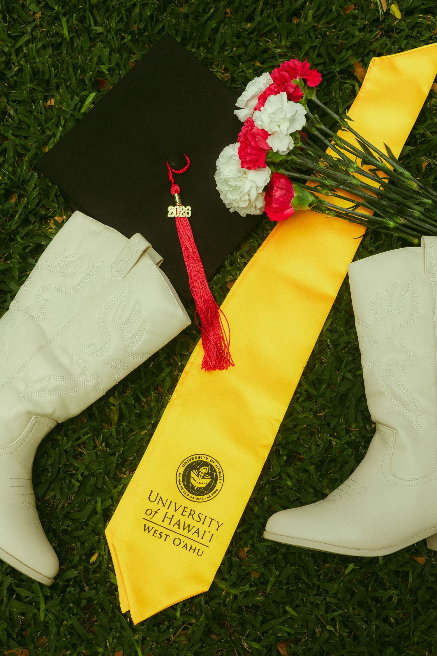 University of Hawaii West Oahu graduation cap with yellow sash, white boots, and flowers on grass.