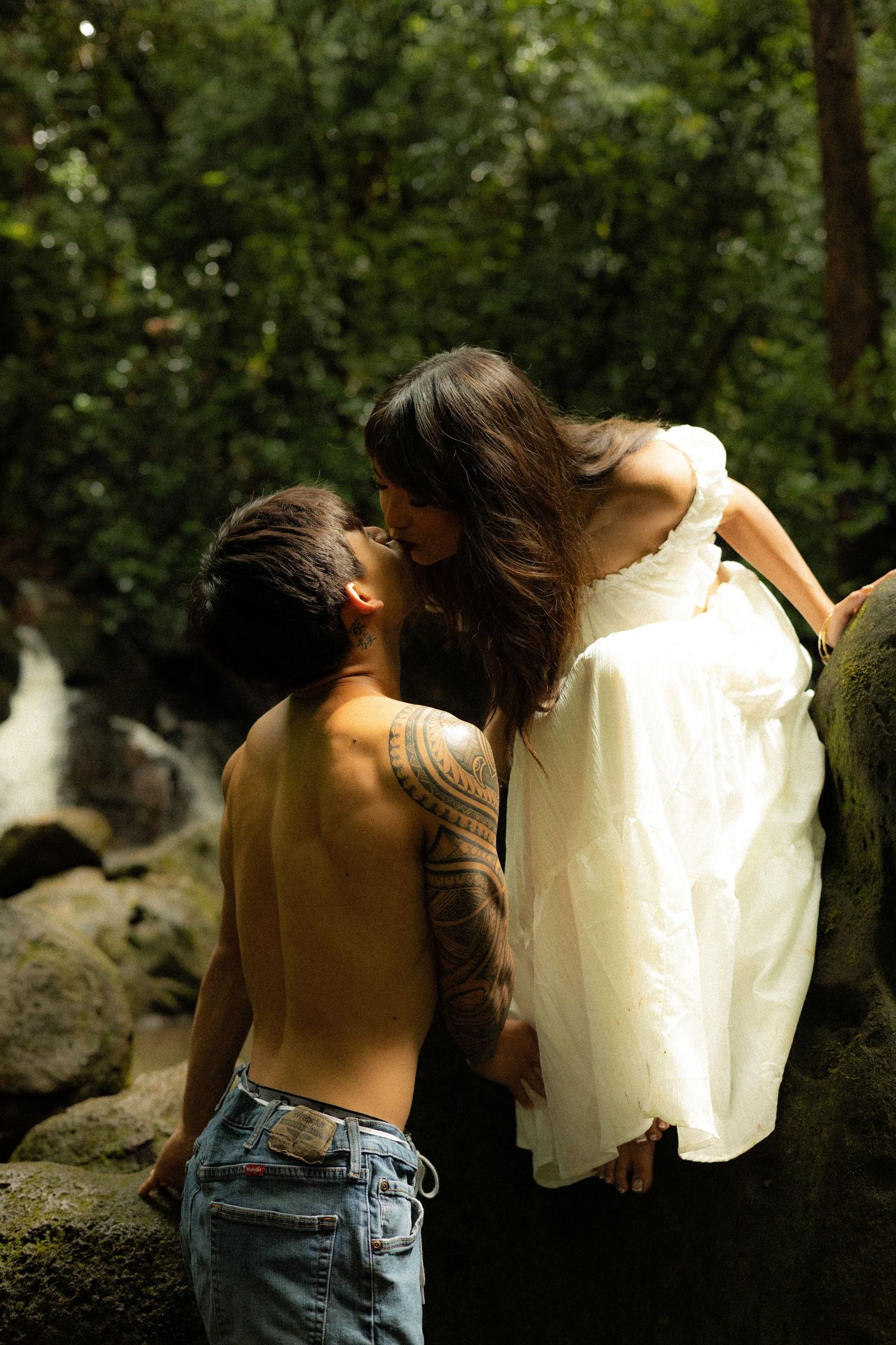 Tattooed shirtless man kissing woman in white dress on mossy rocks near waterfall in lush forest.