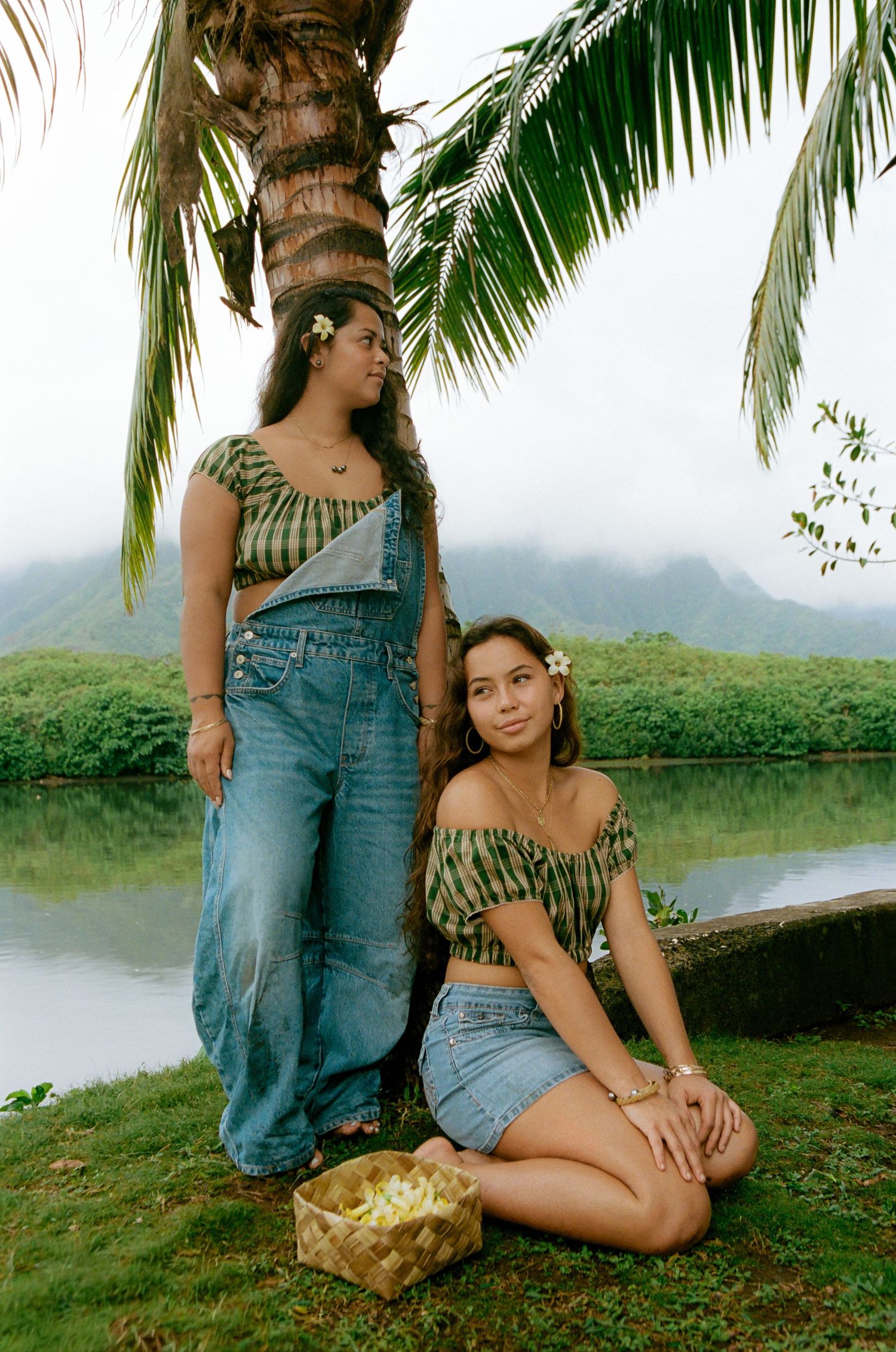 Two women in matching tropical print tops pose under a palm tree by a serene lake with lush green mountains in Hawaii.