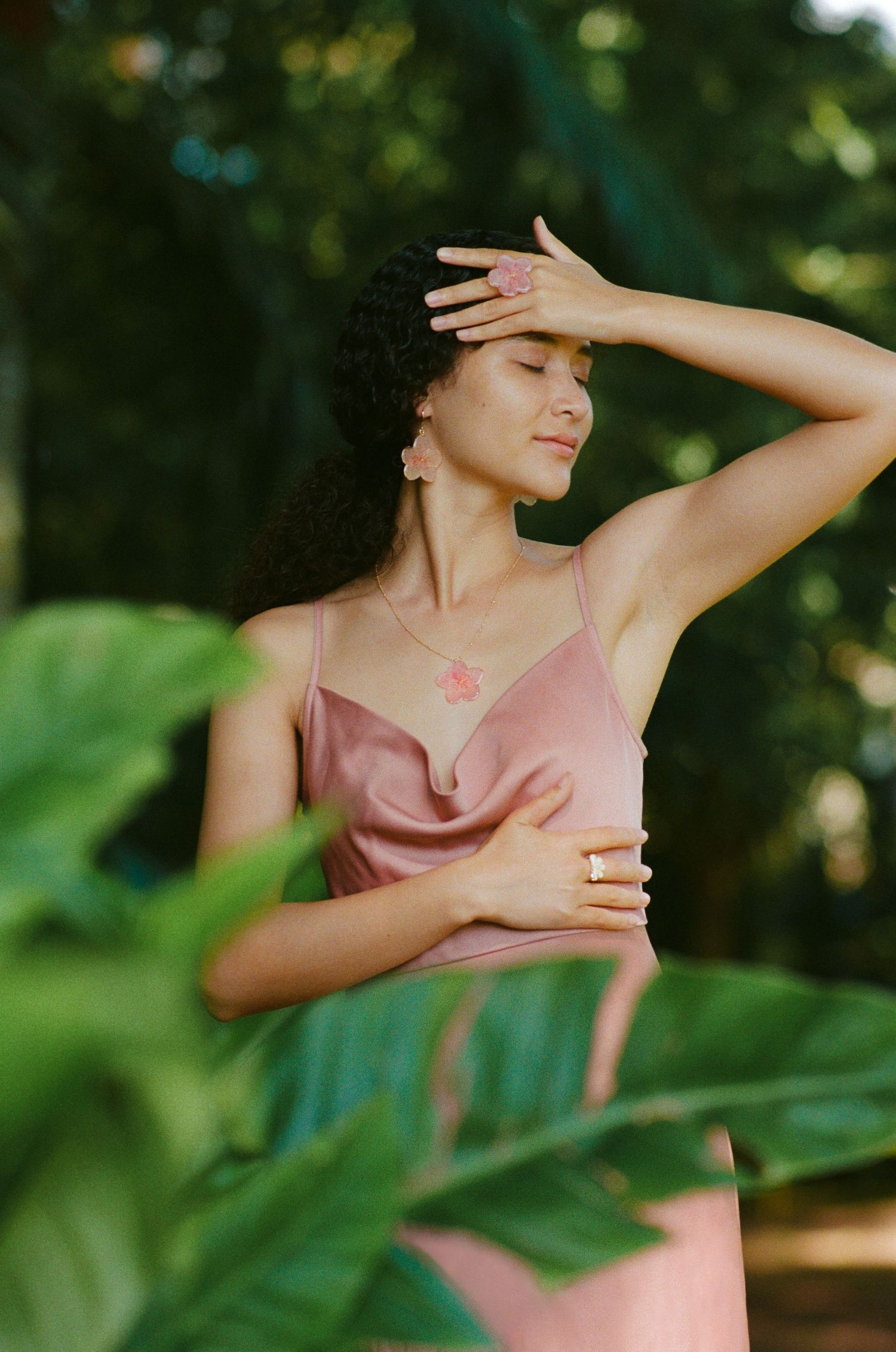 Woman in pink satin slip dress with flower necklace posing among tropical green leaves outdoors.