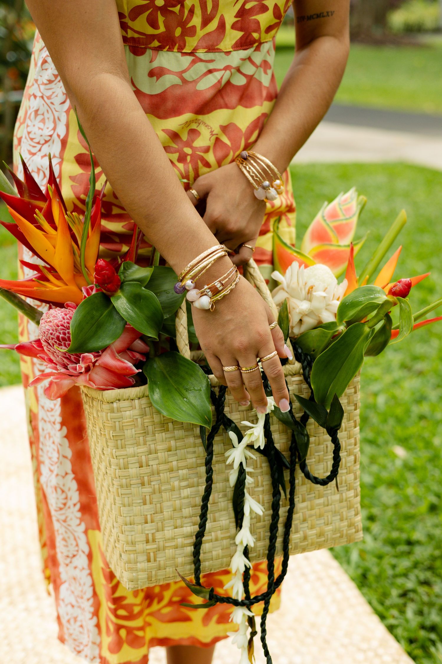Woman in tropical print dress holding woven tote bag filled with bird of paradise flowers and leis.