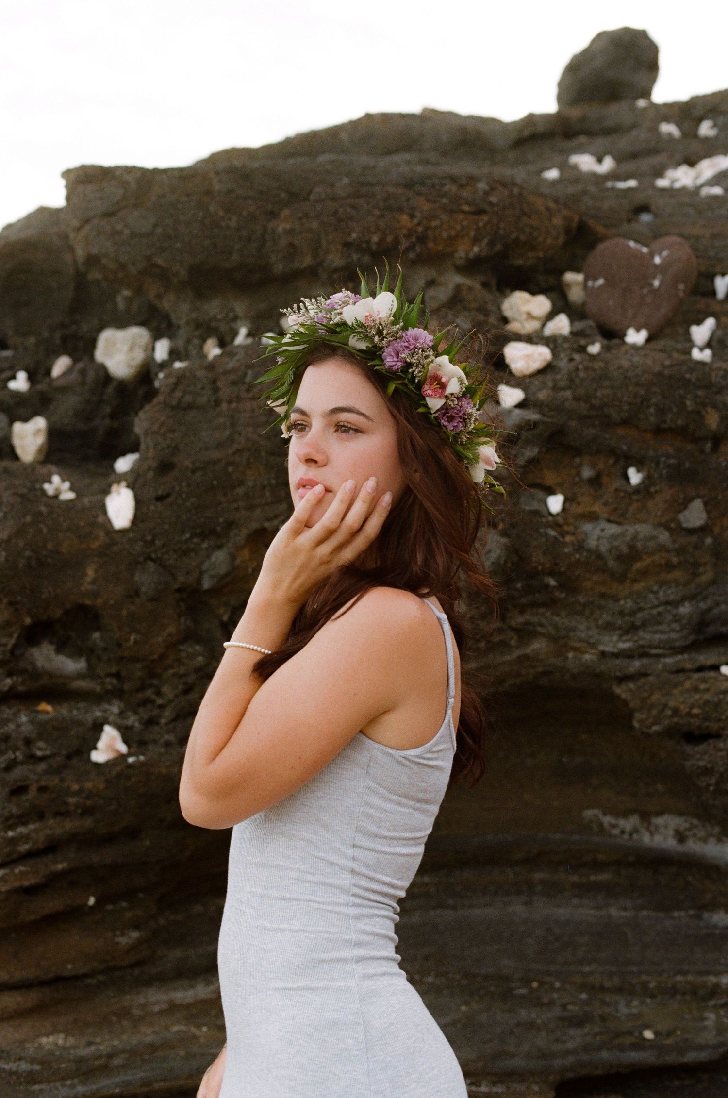 Brunette woman wearing a floral crown and gray dress posing near dark volcanic rocks at the beach.