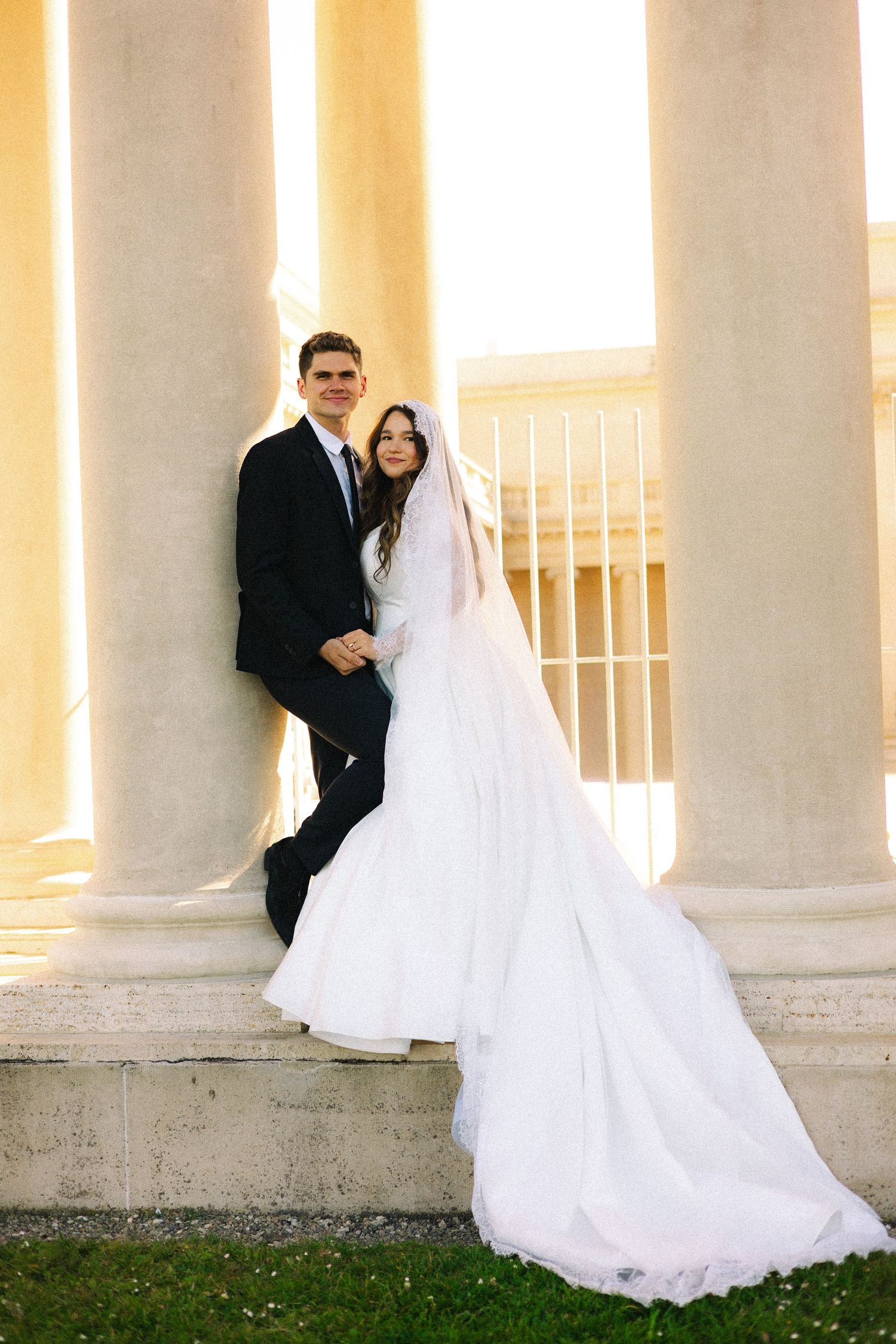 Bride in white gown with veil and groom in black suit pose by white columns at outdoor wedding venue.