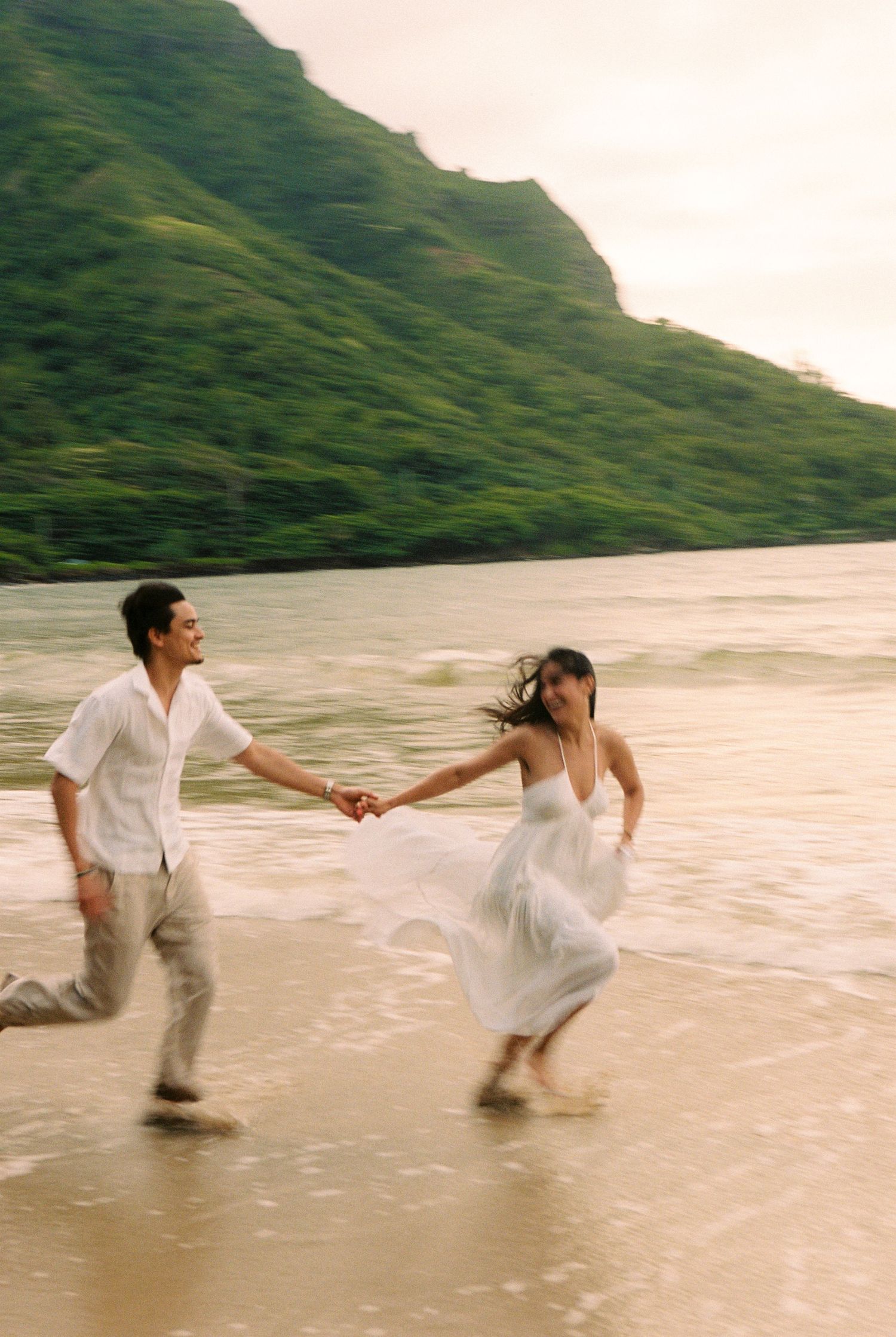 Couple in white outfits running hand-in-hand on a tropical beach with lush green mountains in the background.