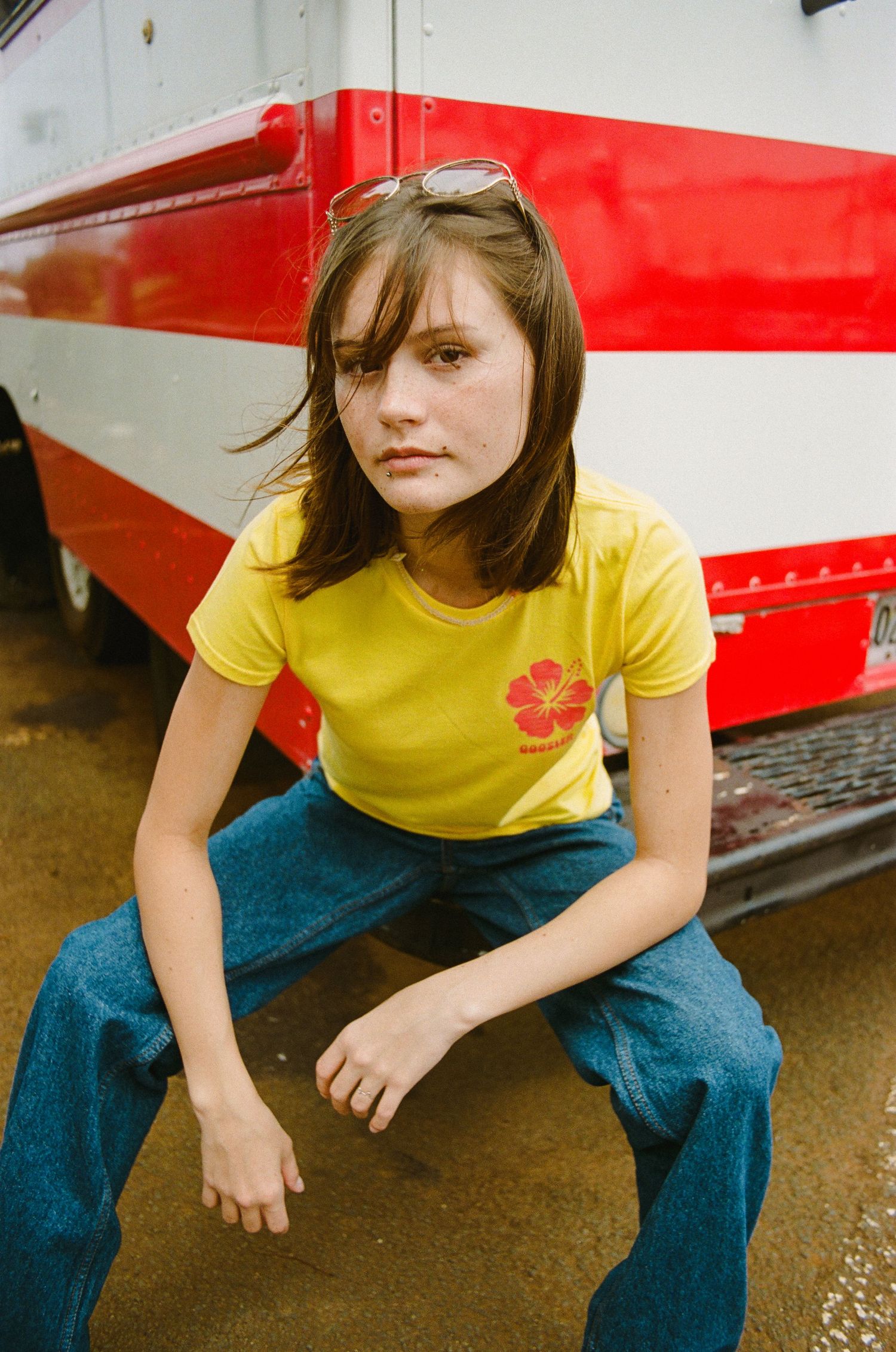 Young woman in yellow hibiscus graphic tee and blue jeans sitting in front of red and white bus.