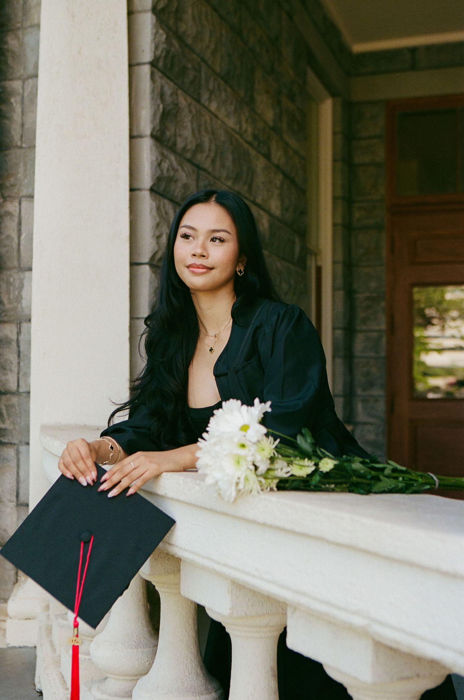 Graduate in black cap and gown holding mortarboard and white flowers on a stone building porch.