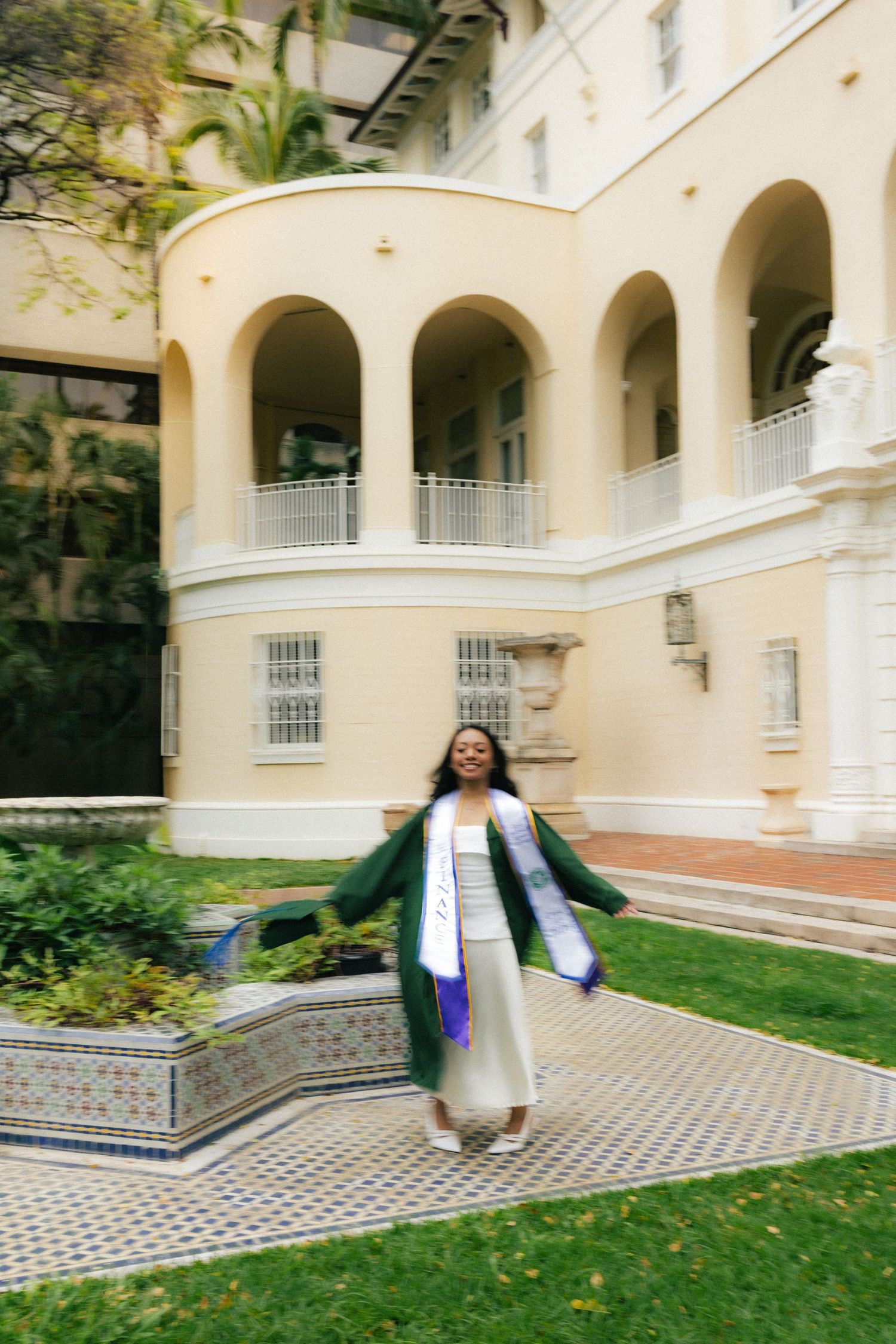 Graduate in green cap and gown spinning joyfully in courtyard of elegant yellow arched building.