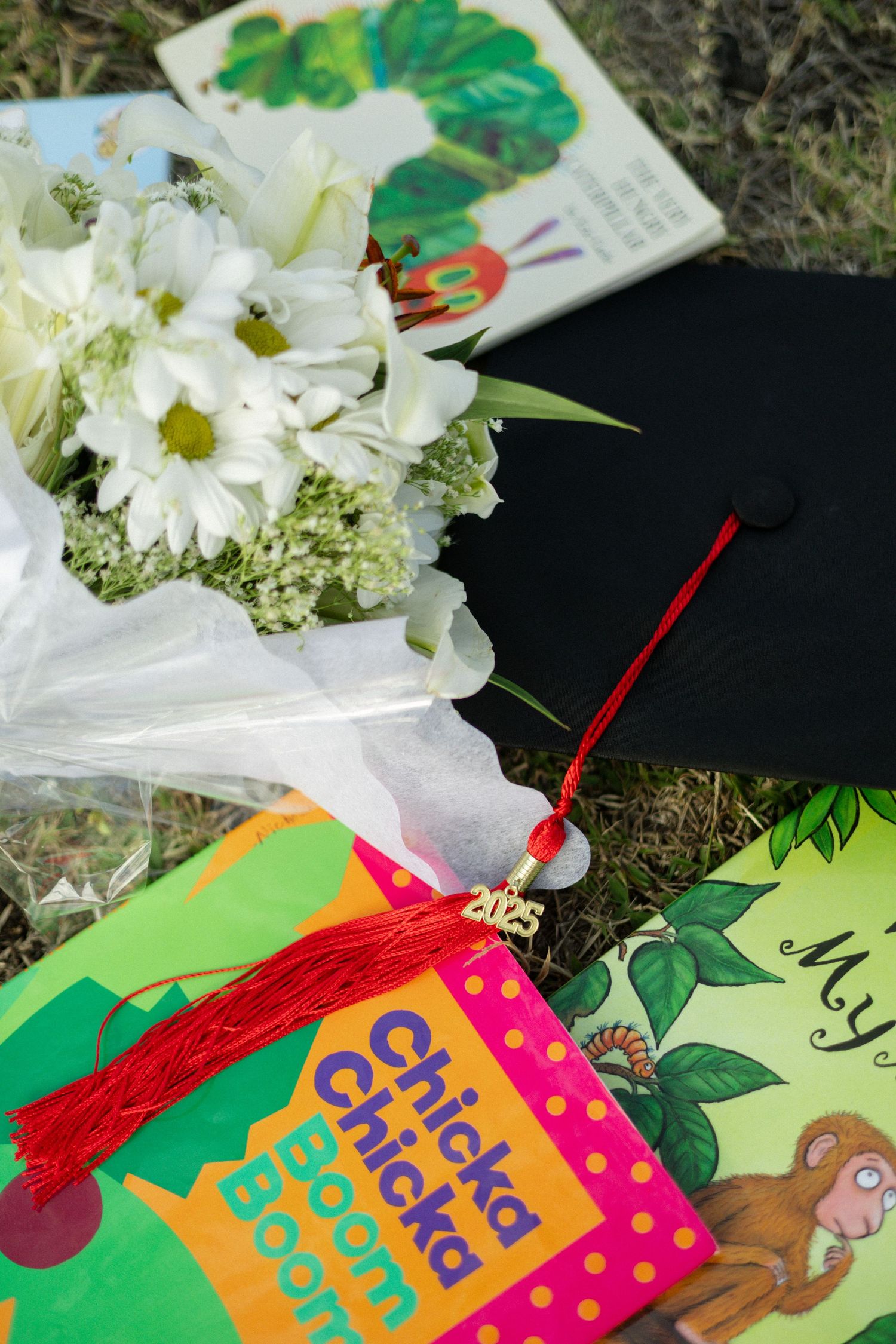 Graduation cap with red tassel, white flowers, and childrens books on grass for 2023 ceremony.
