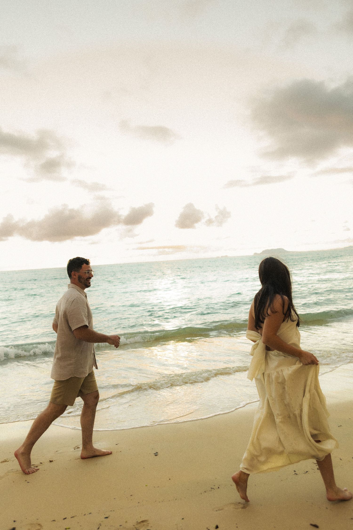 Couple walking barefoot along sandy beach shoreline at sunset with cloudy sky and ocean waves.
