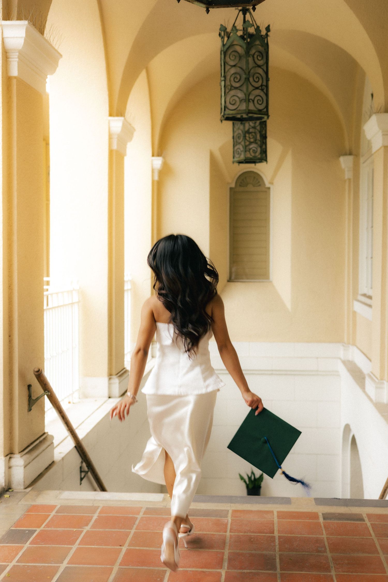Graduate in white dress holding green cap walking through elegant arched hallway from behind.
