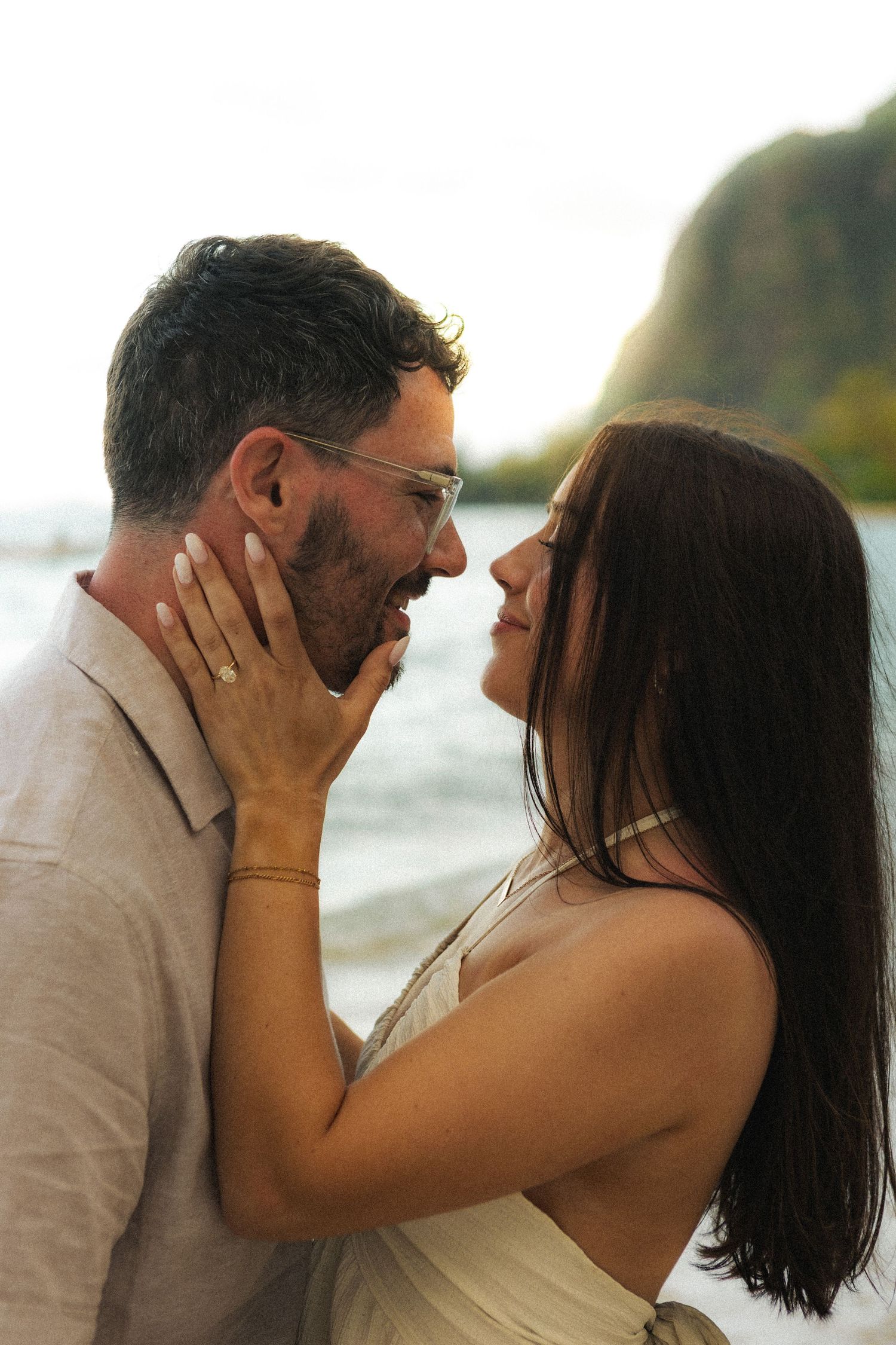 Romantic couple sharing a tender moment on a tropical beach, woman touching man's face with engagement ring visible.