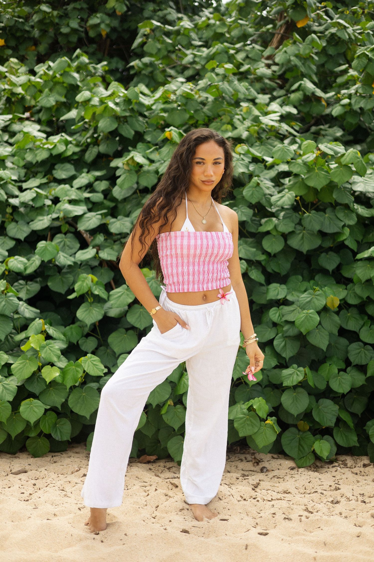 Woman in pink gingham halter top and white pants standing before lush green foliage.
