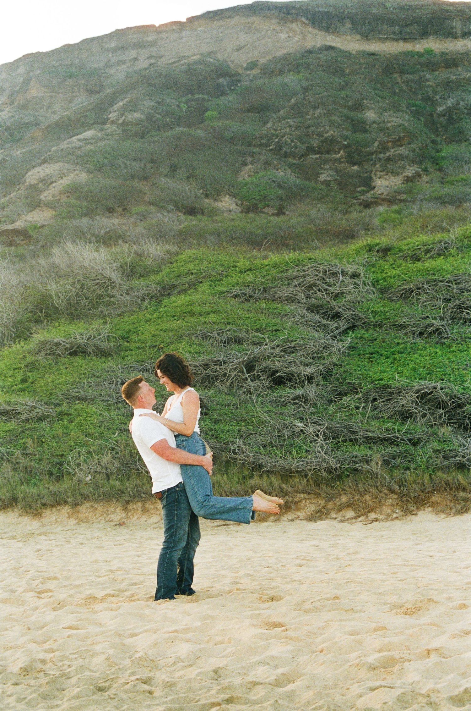 A man lifts his partner joyfully on a sandy beach with lush green hills in the background.