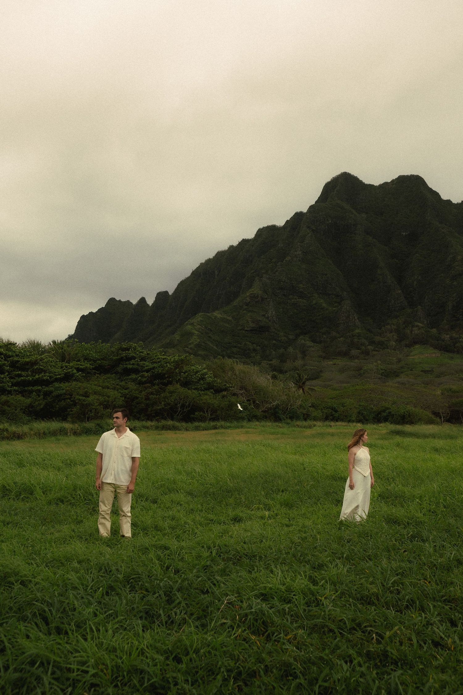 Couple in white standing apart in lush green field with dramatic mountain backdrop in Hawaii.