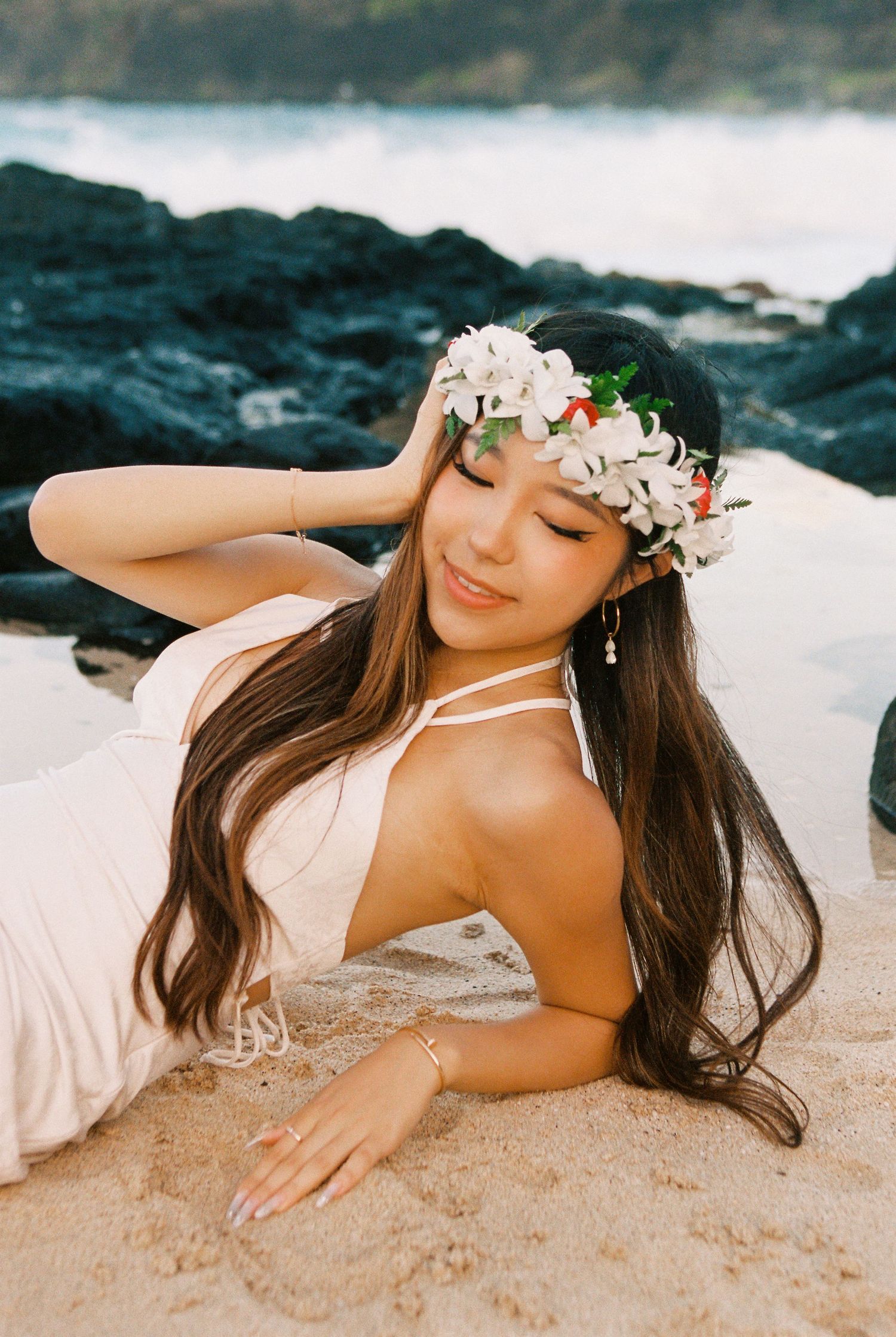 Woman in white dress and floral crown poses gracefully on sandy beach with dark rocks in background.