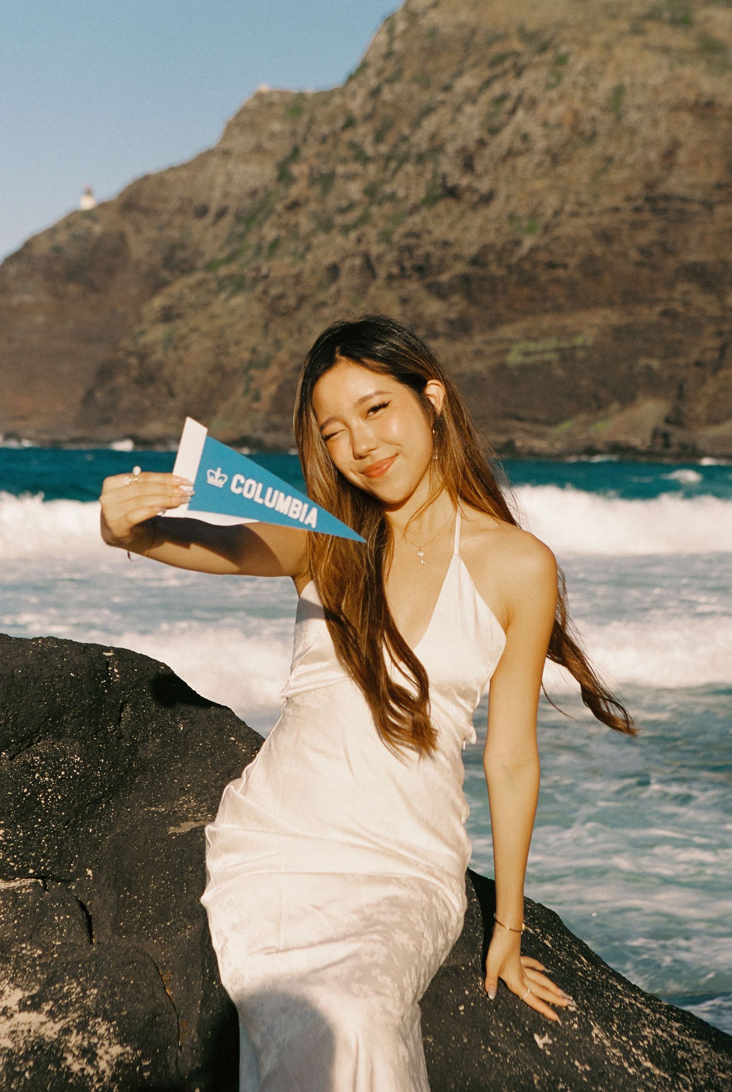 Young woman in white dress holding a Columbia University pennant flag, sitting on rocks by the ocean.